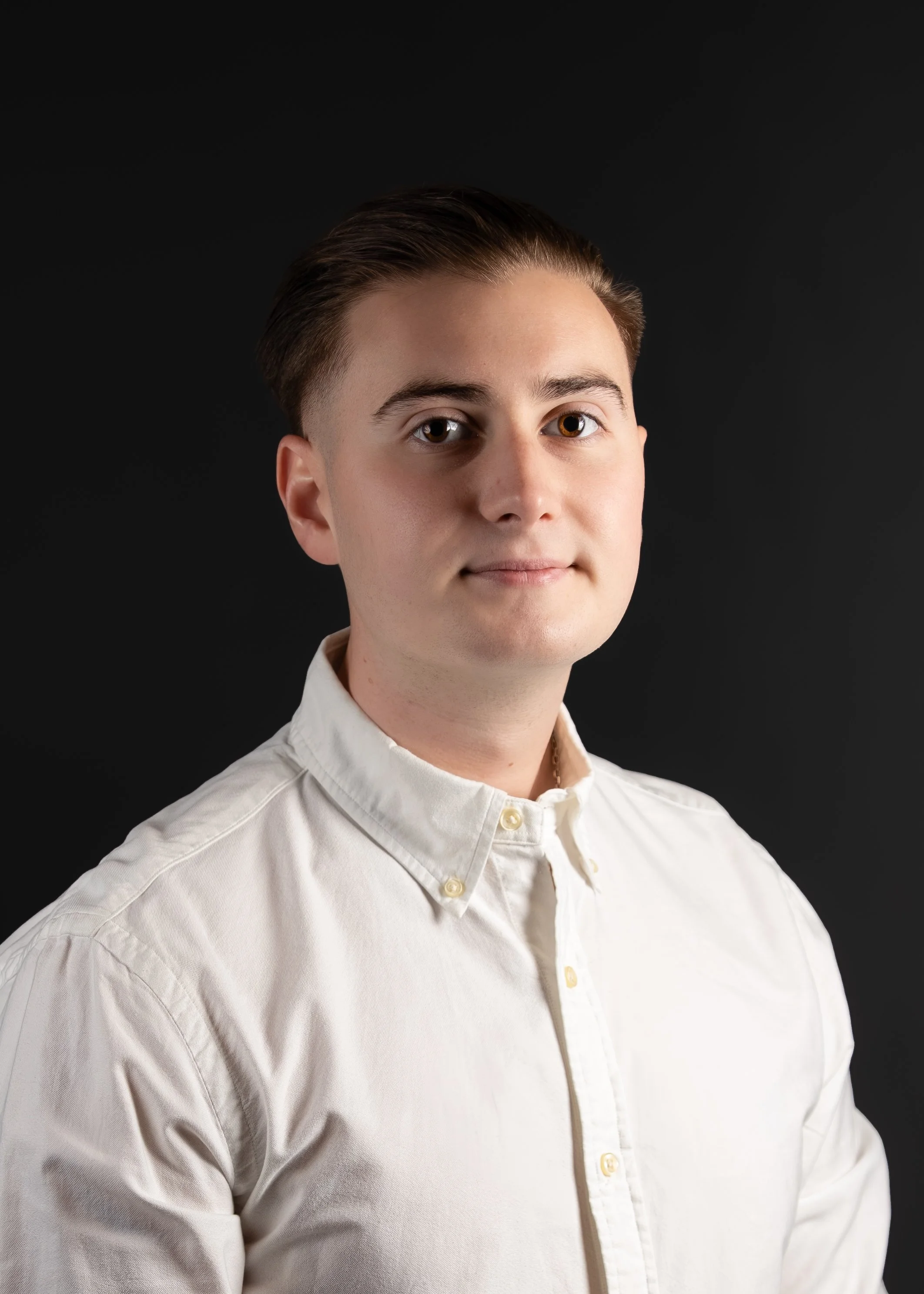A young man with short brown hair, fair skin, wearing a white button-up shirt, against a solid black background.