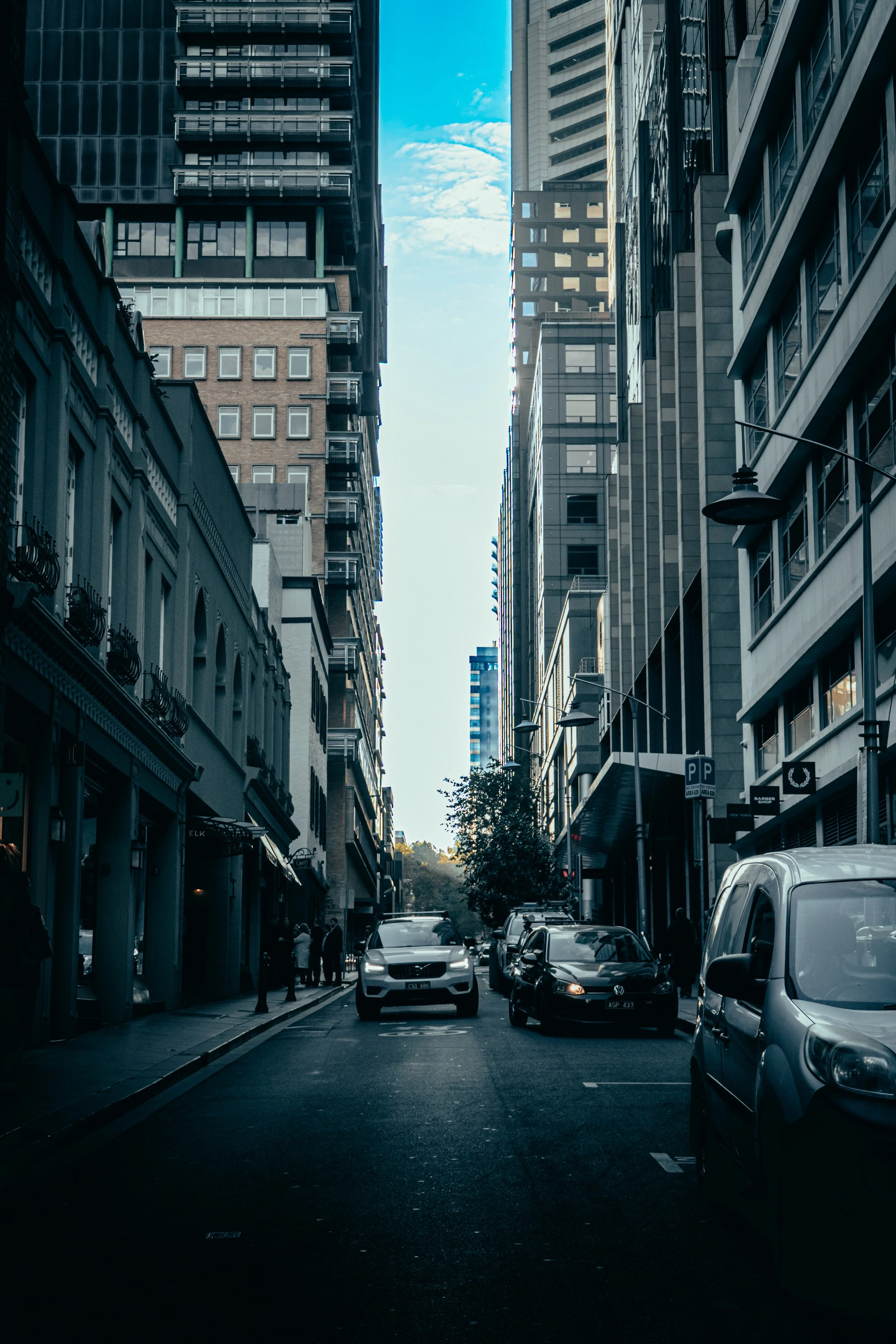 A city street lined with tall modern buildings on both sides, with parked cars along the curb and a few pedestrians on the sidewalk, under a partly cloudy sky.
