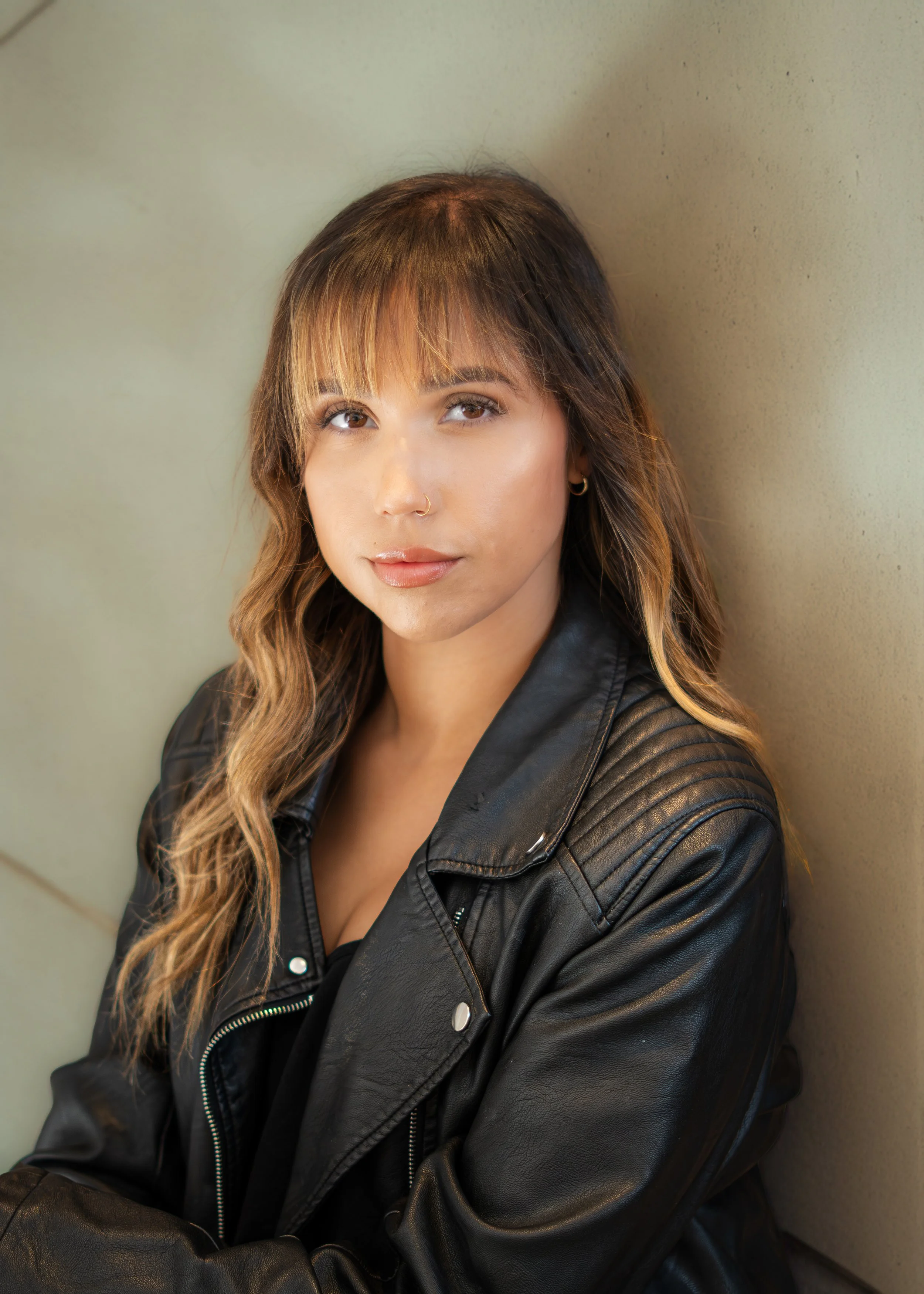 A young woman with light skin and long wavy hair, wearing a black leather jacket, leaning against a beige wall.