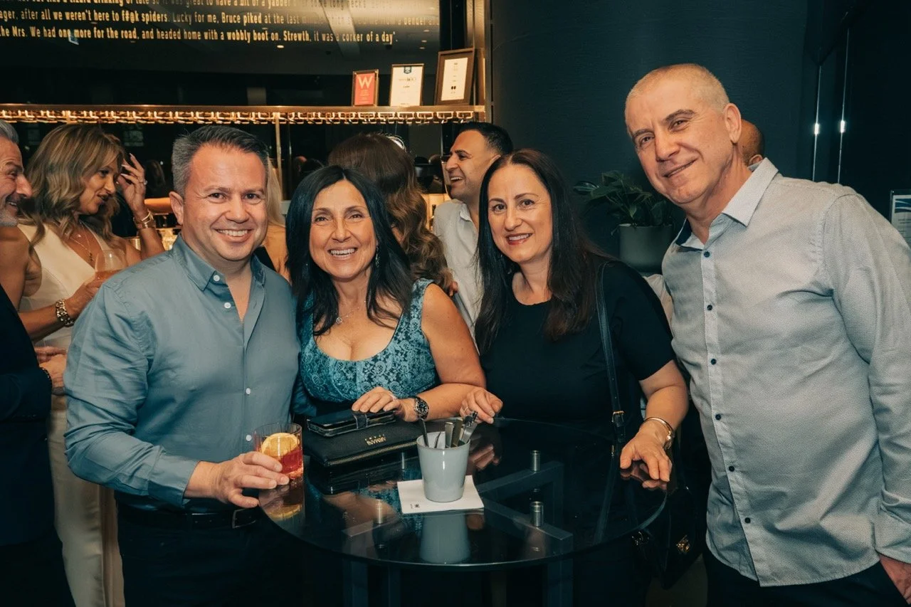 Group of people at a social gathering, smiling and enjoying drinks in a modern indoor setting.
