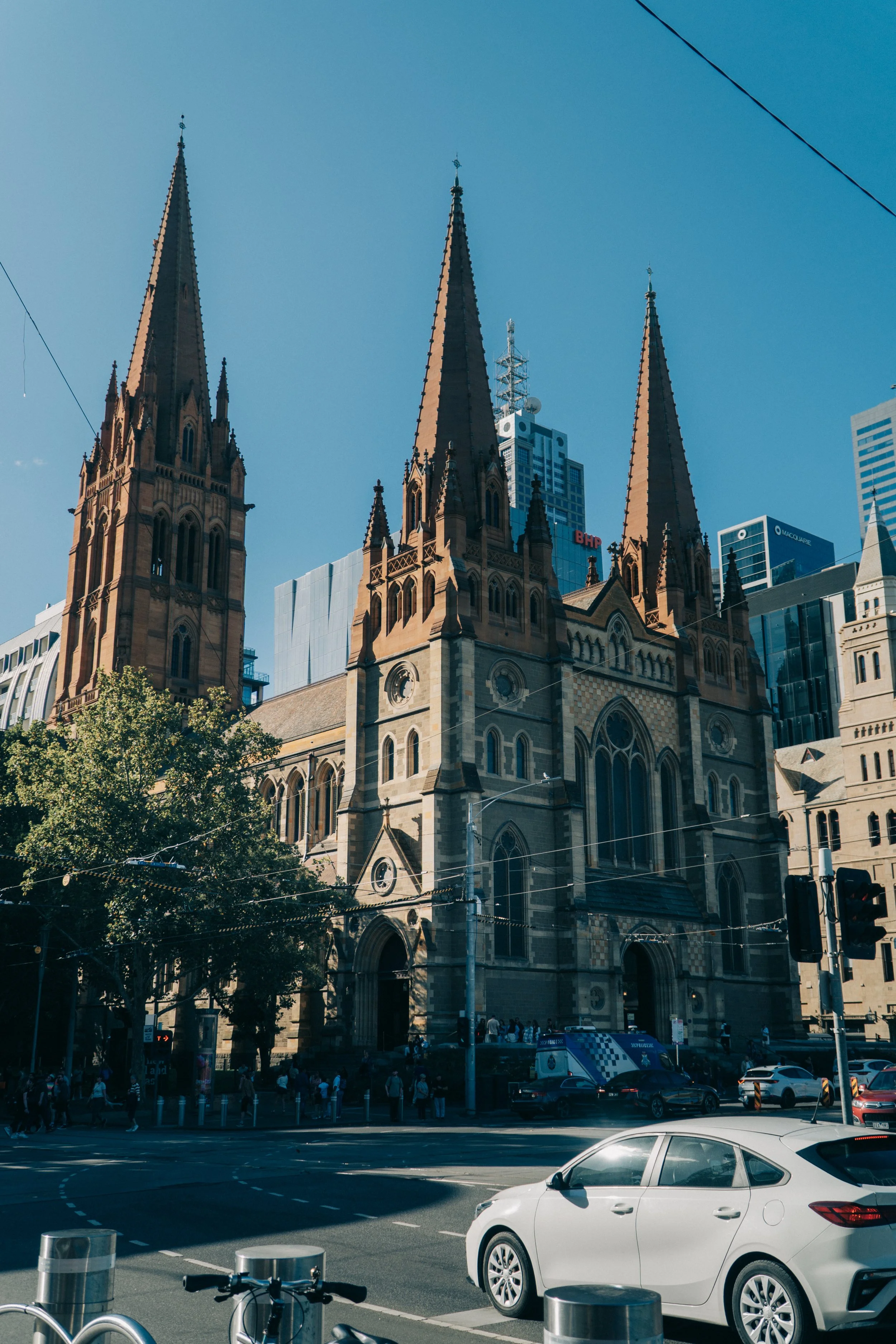 A large Gothic-style church with three tall spires, surrounded by modern high-rise buildings, trees, and a busy street with cars and pedestrians.