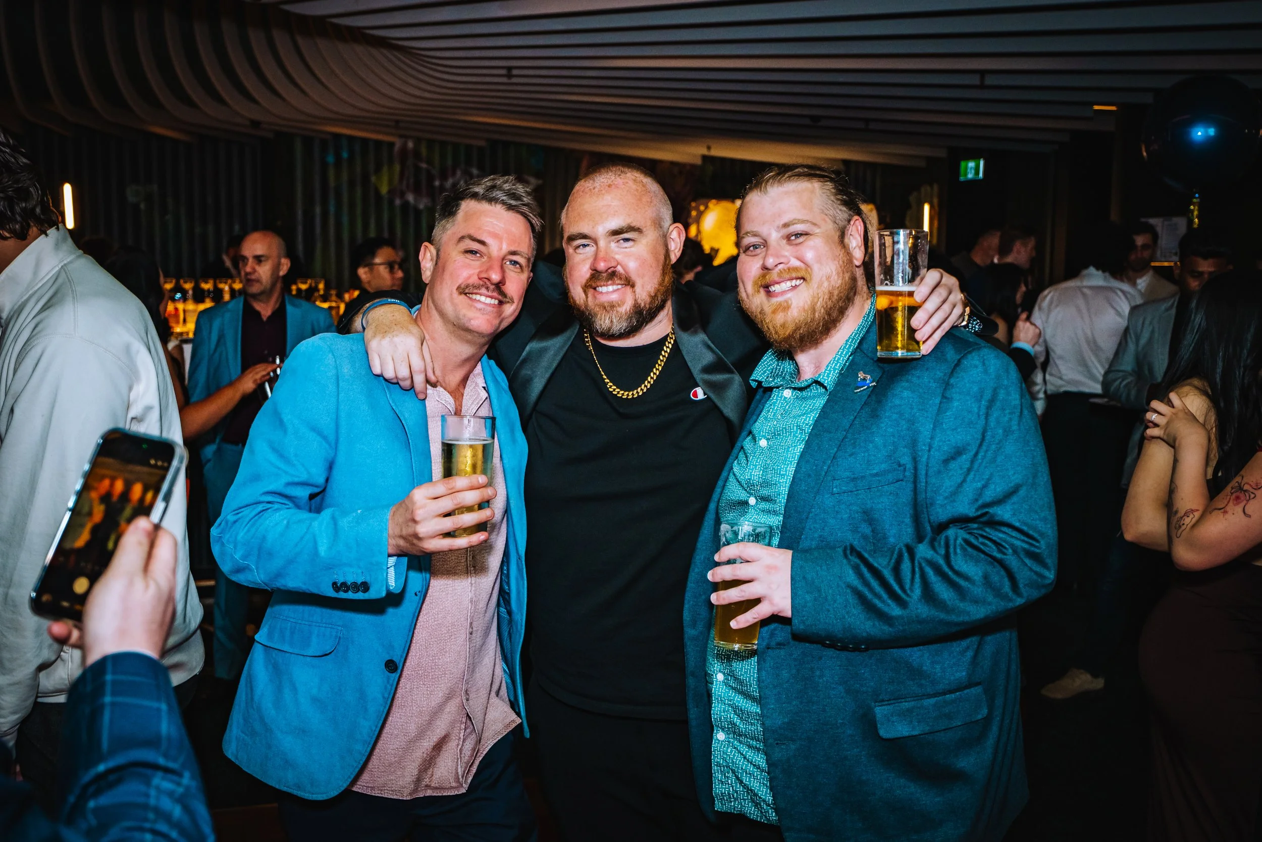 Three men smiling and holding drinks at a party or bar, with others in the background.