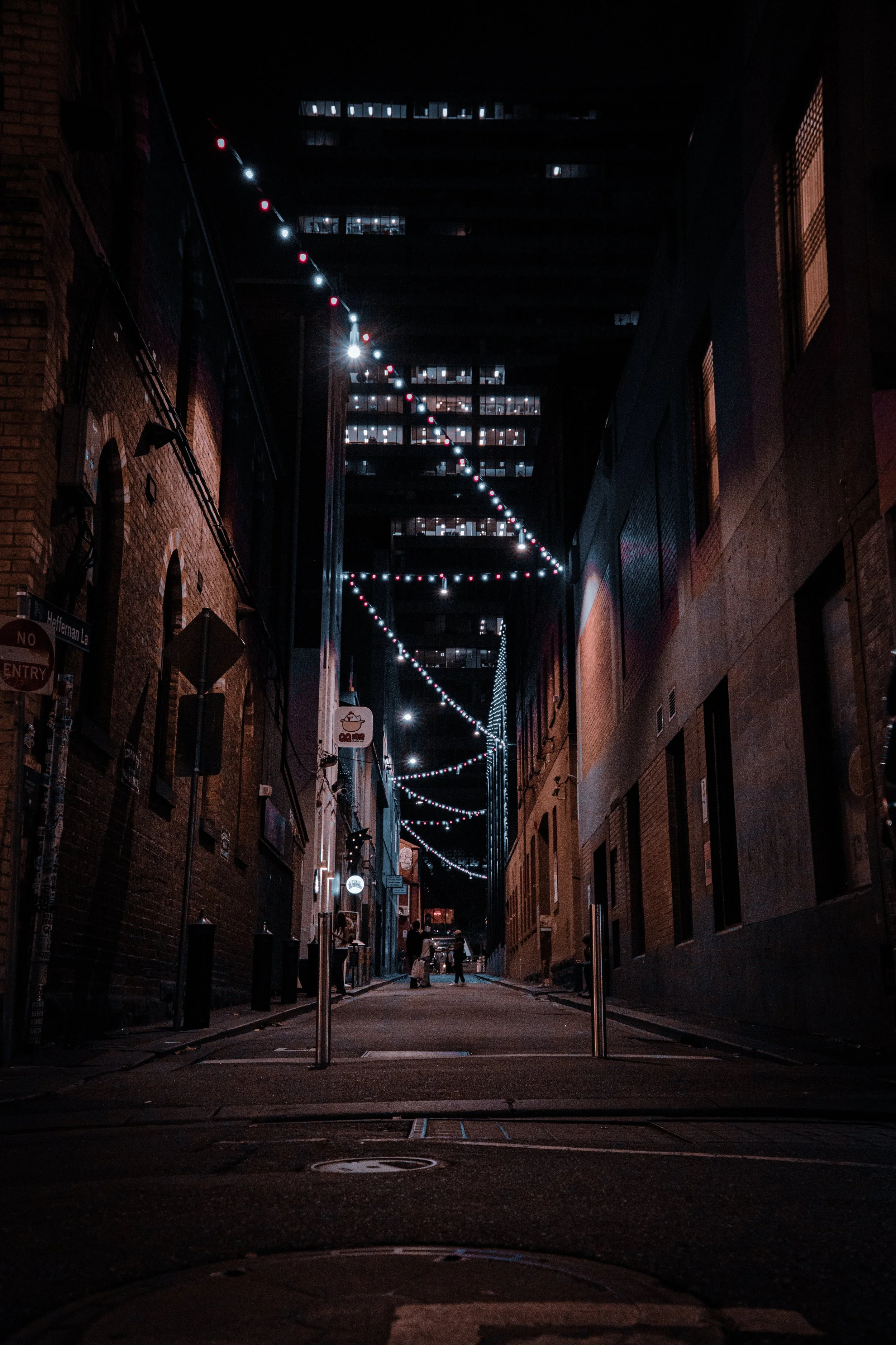 Nighttime alleyway with string lights hanging overhead, surrounded by tall brick and concrete buildings, with a few people walking in the distance.