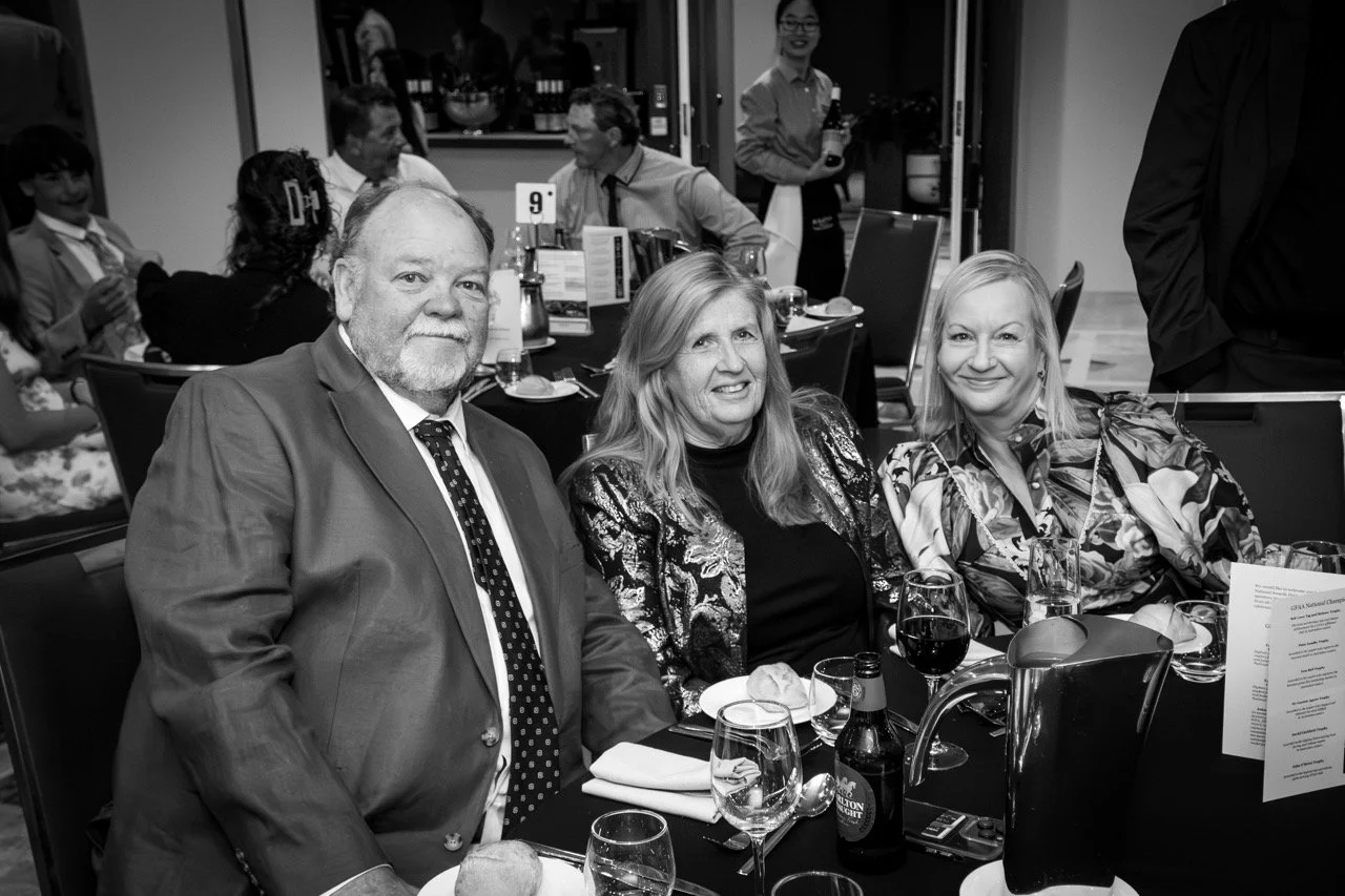 Group of people sitting at a banquet table during a formal event, with three people in the foreground smiling at the camera, two men and one woman, surrounded by glasses and table settings.