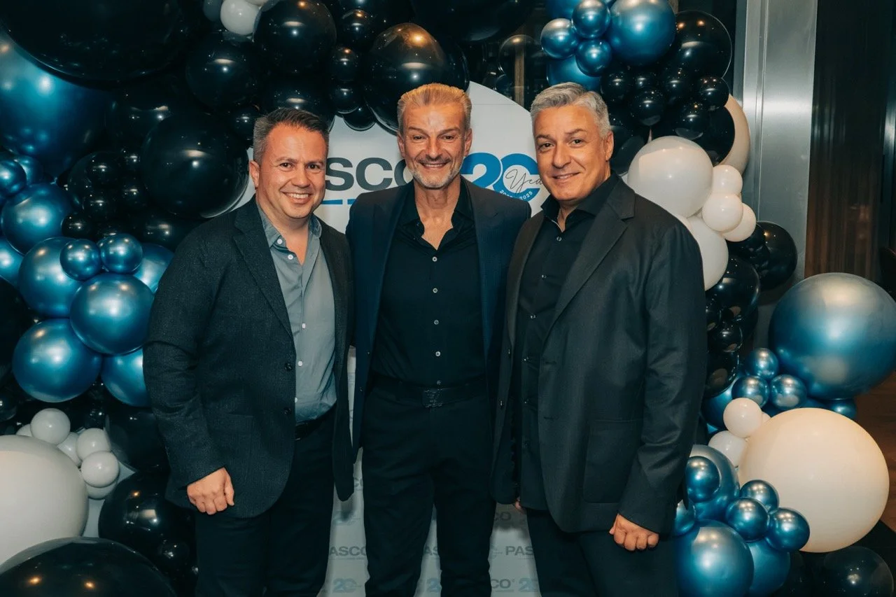 Three men in suits smiling at an event with a backdrop and a balloon decoration with black, white, and blue balloons celebrating a 20th anniversary.