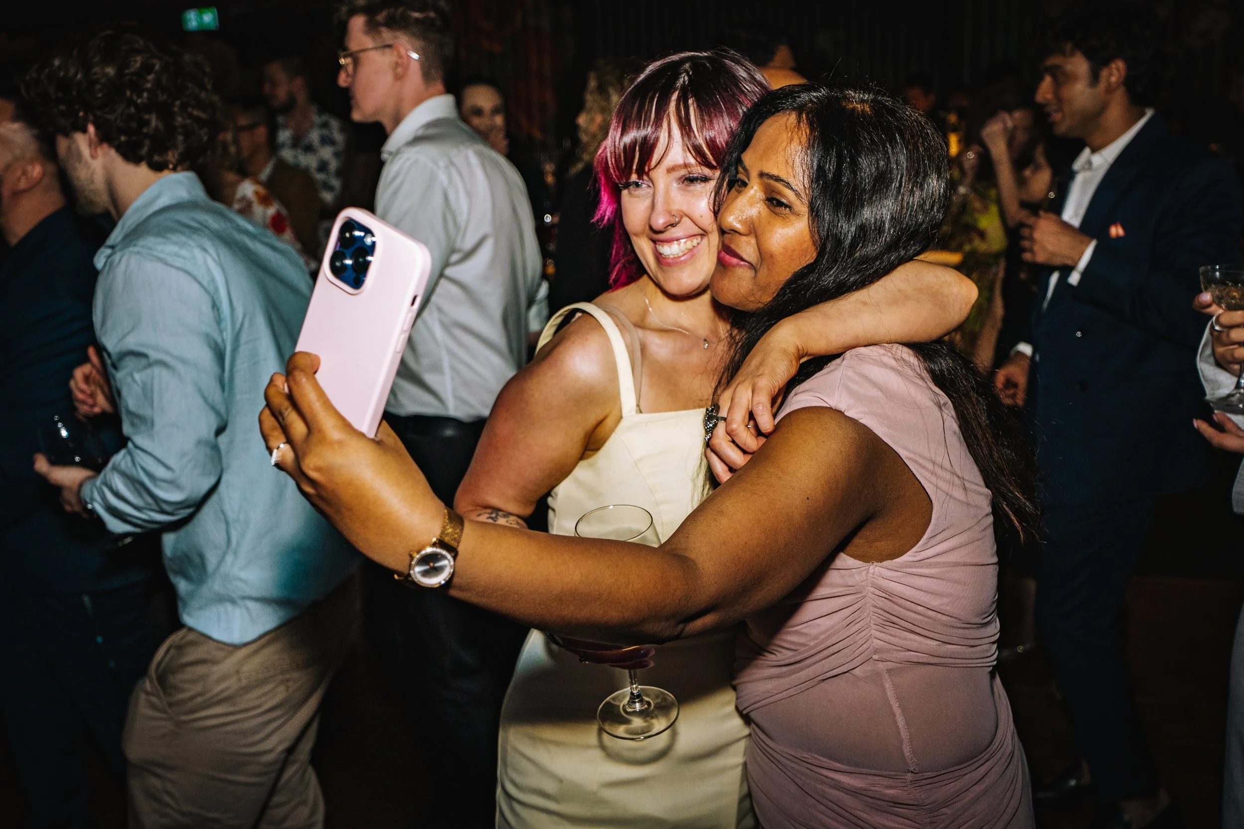 Two women hugging and taking a selfie at a lively social event with other people mingling in the background.