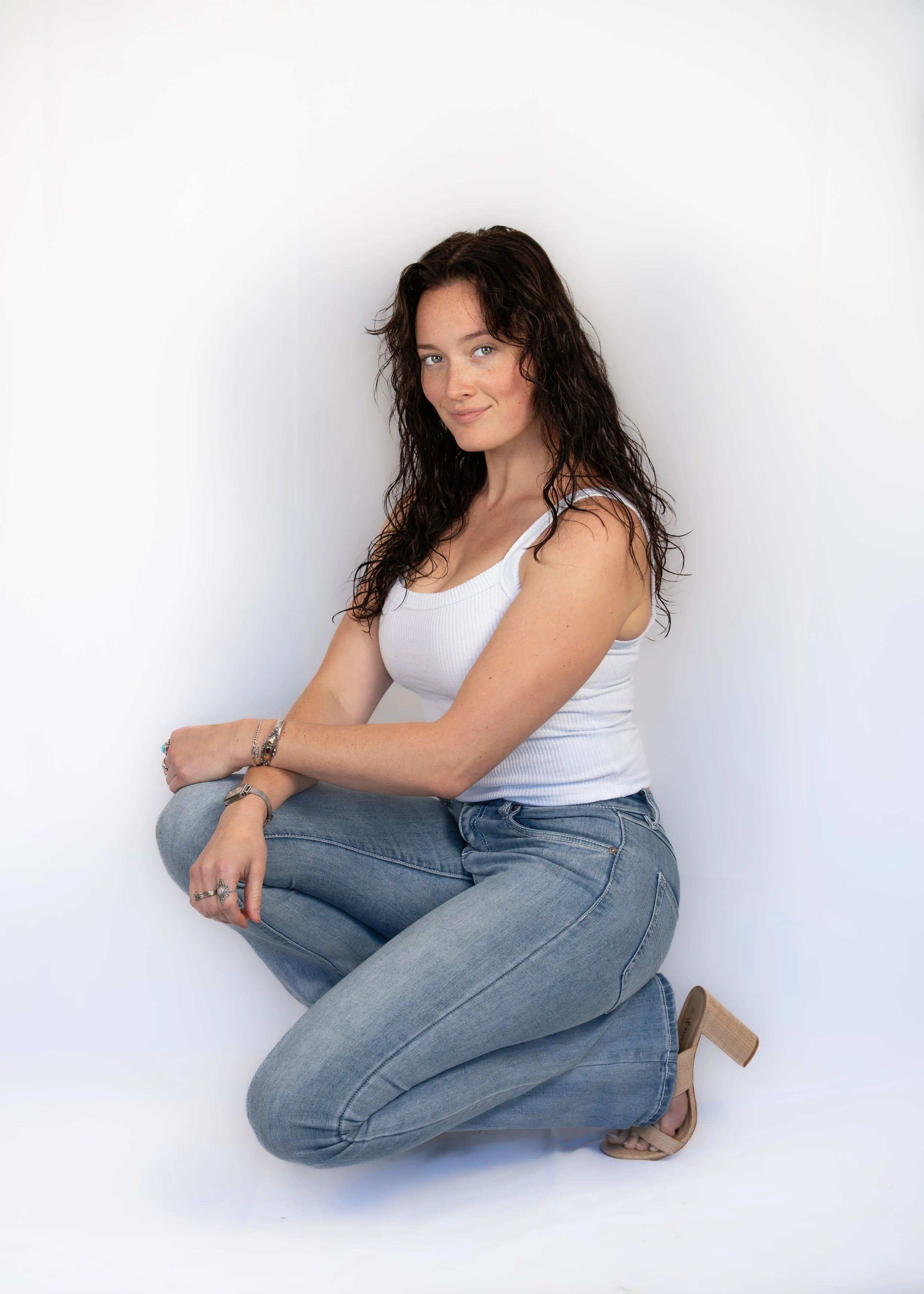Young woman with curly brown hair and light skin, wearing a white tank top and light blue jeans, sitting against a white background in a relaxed pose with a gentle smile.