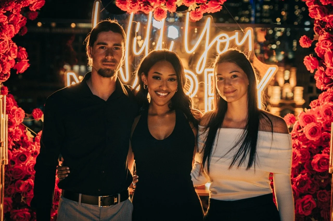 Three friends standing together at night, smiling, with a floral backdrop and neon sign that says "Until You" in the background.