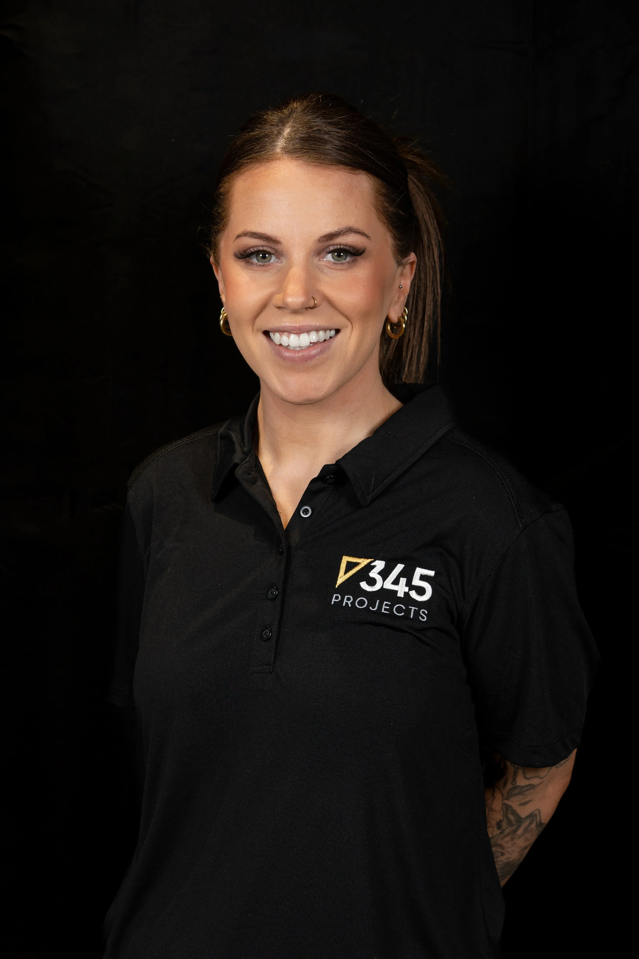 A woman with brown hair, tattoos, and hoop earrings is smiling and wearing a black polo shirt with the logo '345 PROJECTS' on it, standing against a black background.