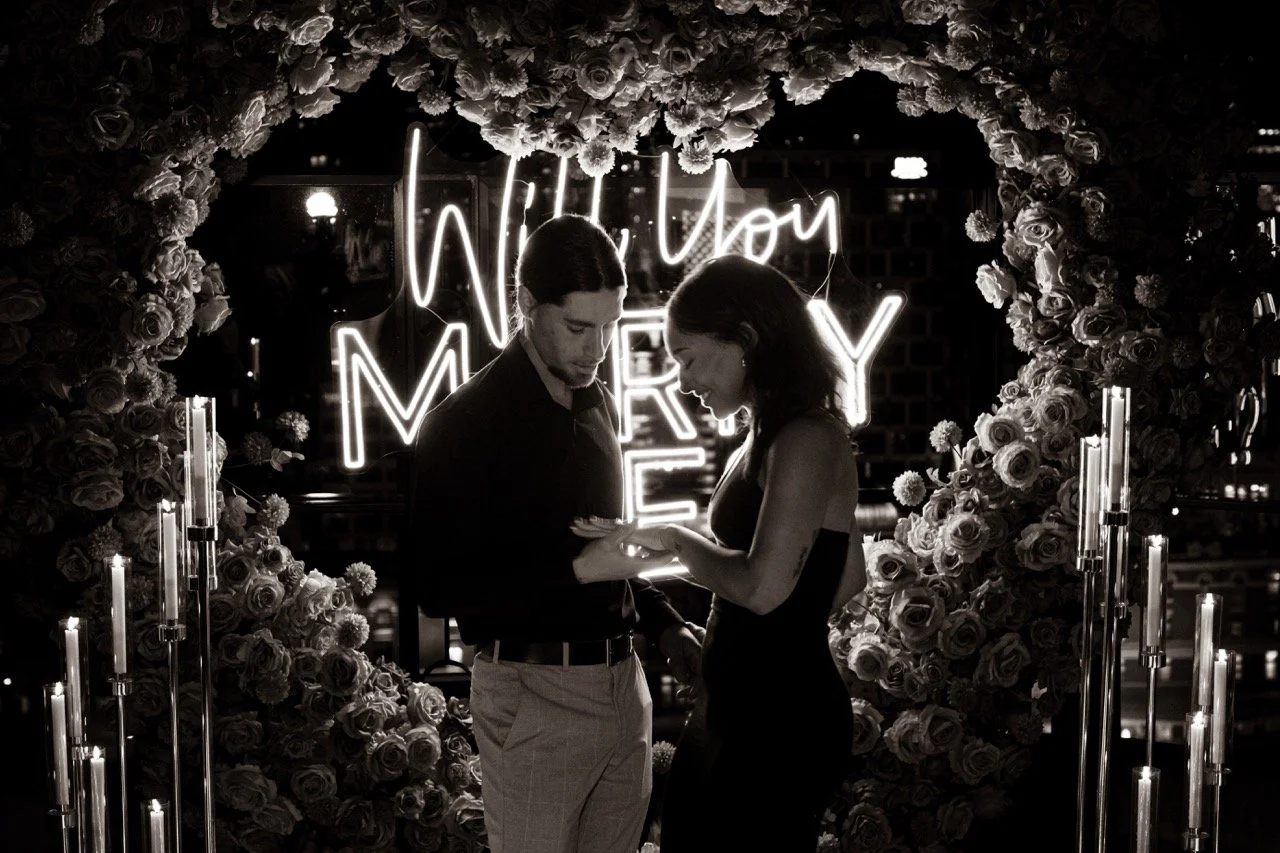 A couple on a romantic date, with a heart-shaped flower wreath and neon sign in the background that reads 'Will You Marry Me', illuminated in a dark setting.