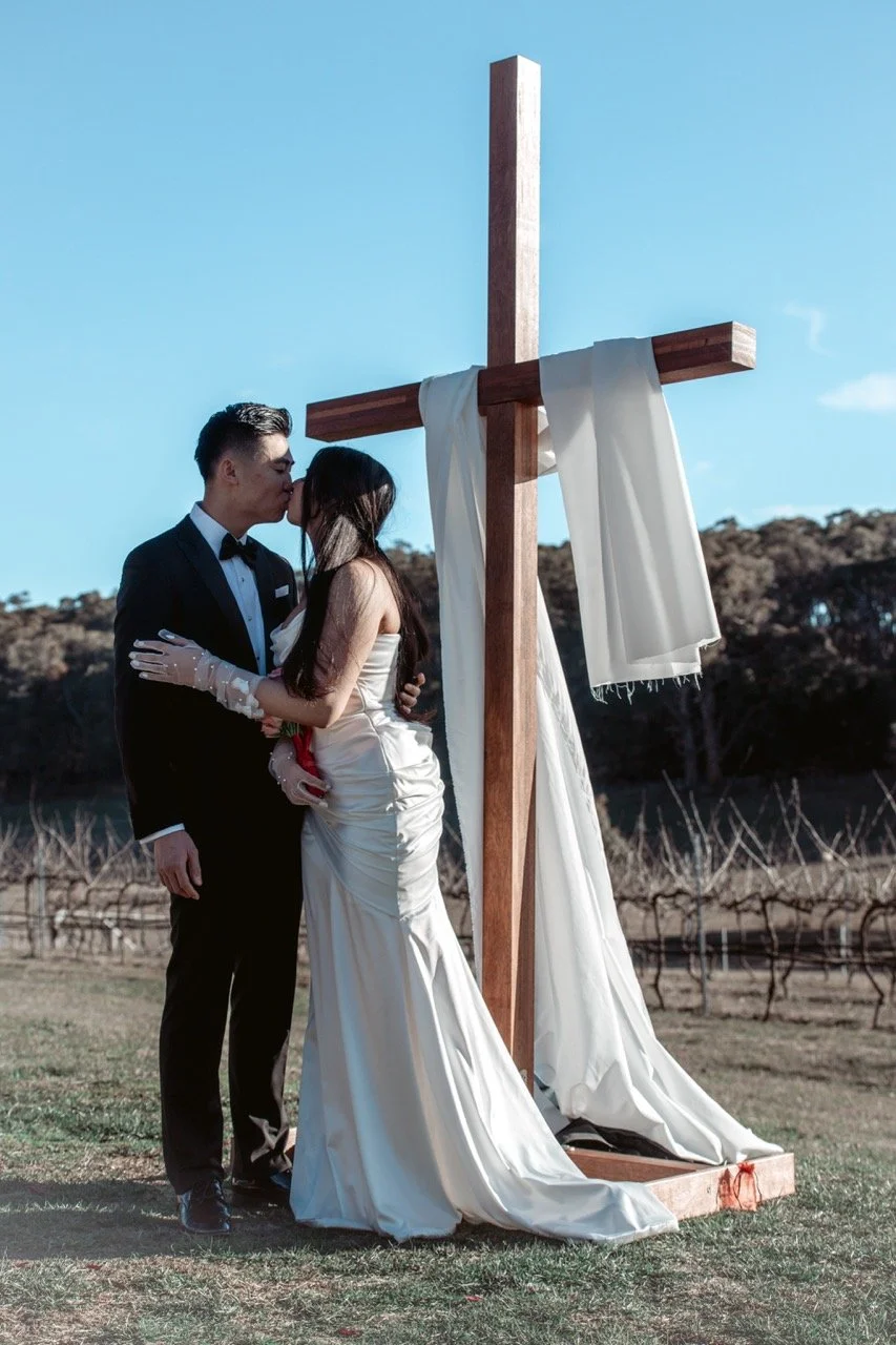 A couple dressed in wedding attire kissing outdoors near a large wooden cross with white fabric draped on it, in a natural setting with a vineyard and trees in the background.