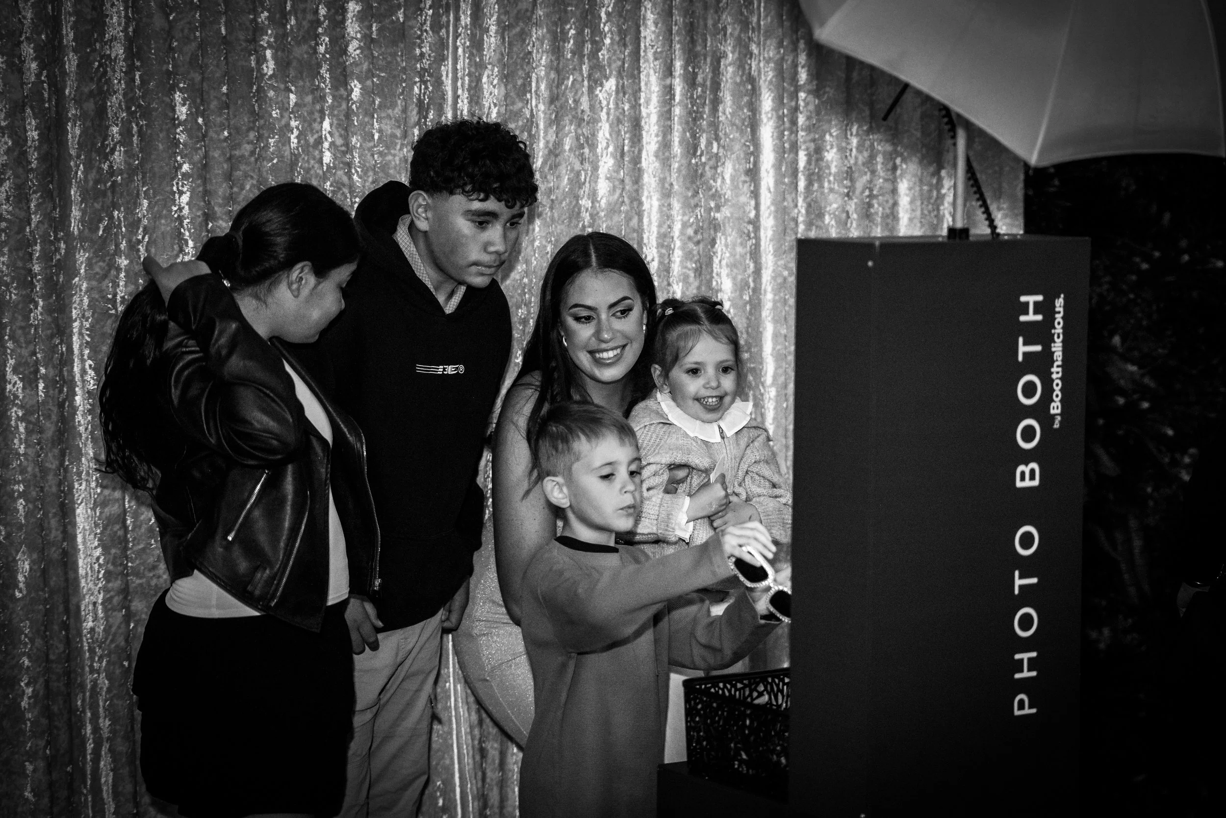 A group of five people, including two children, are gathered in front of a photo booth with a black and white curtain background. One young girl is holding sunglasses, and they are all looking at the photo booth, smiling or with interest.