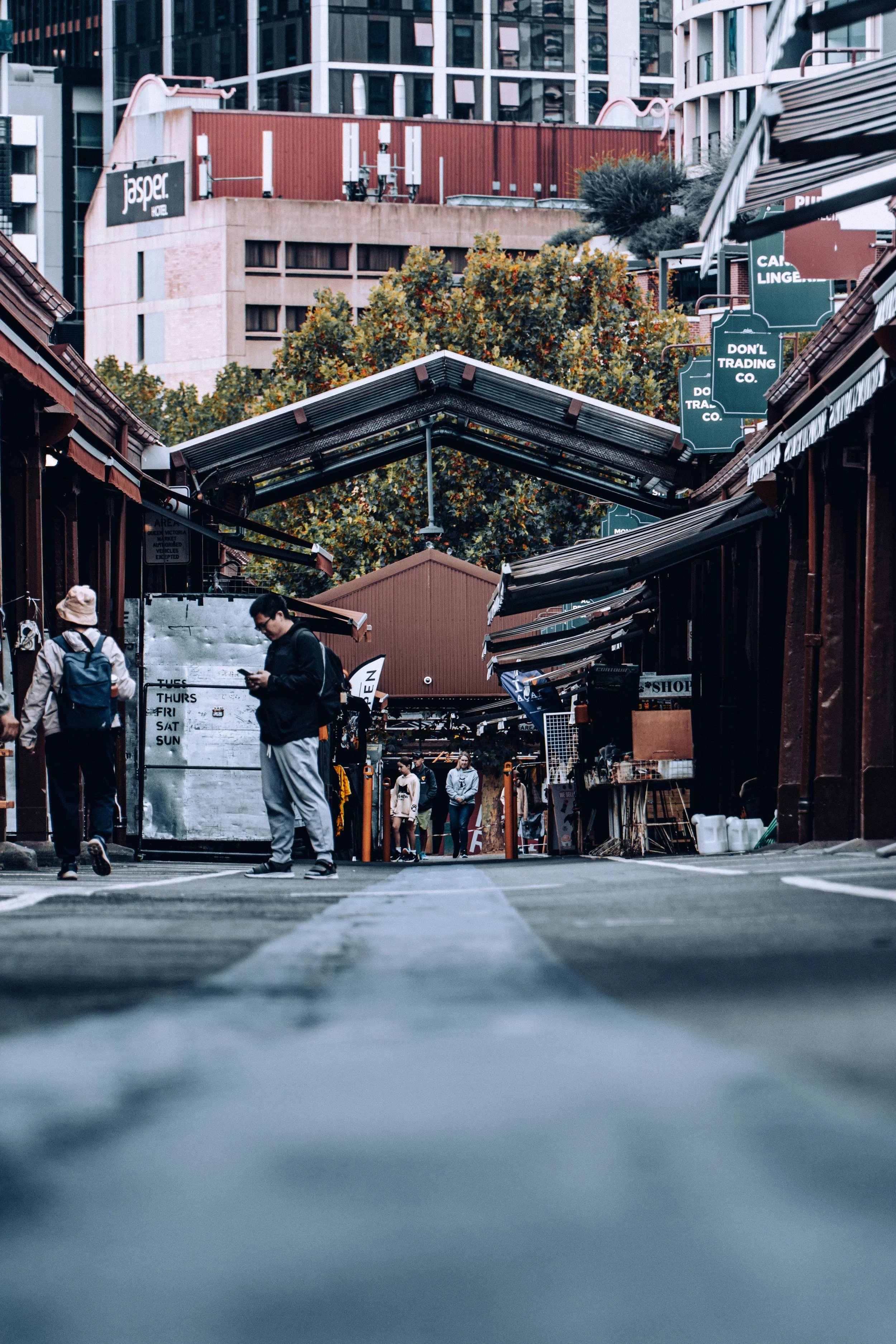 View of a marketplace with shops on either side and people walking, with tall buildings in the background and signs hanging overhead.