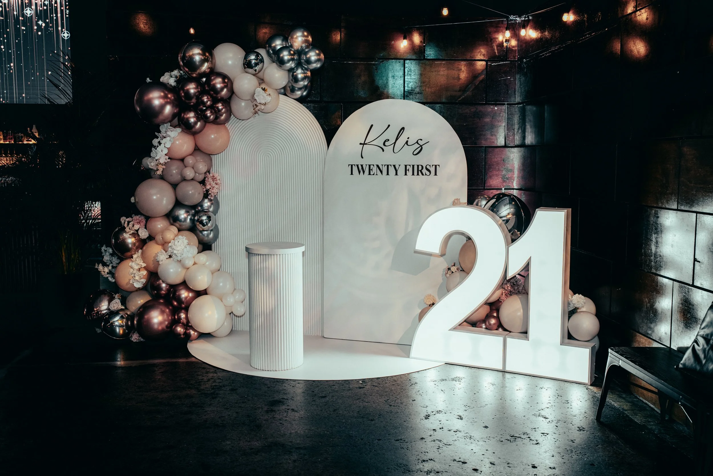 Decorative setup for a 21st birthday celebration featuring a large illuminated '21' sign, a balloon arrangement with pink, white, and metallic balloons, and a backdrop with the name 'Kelis' and text 'Twenty First' against a dark wall.