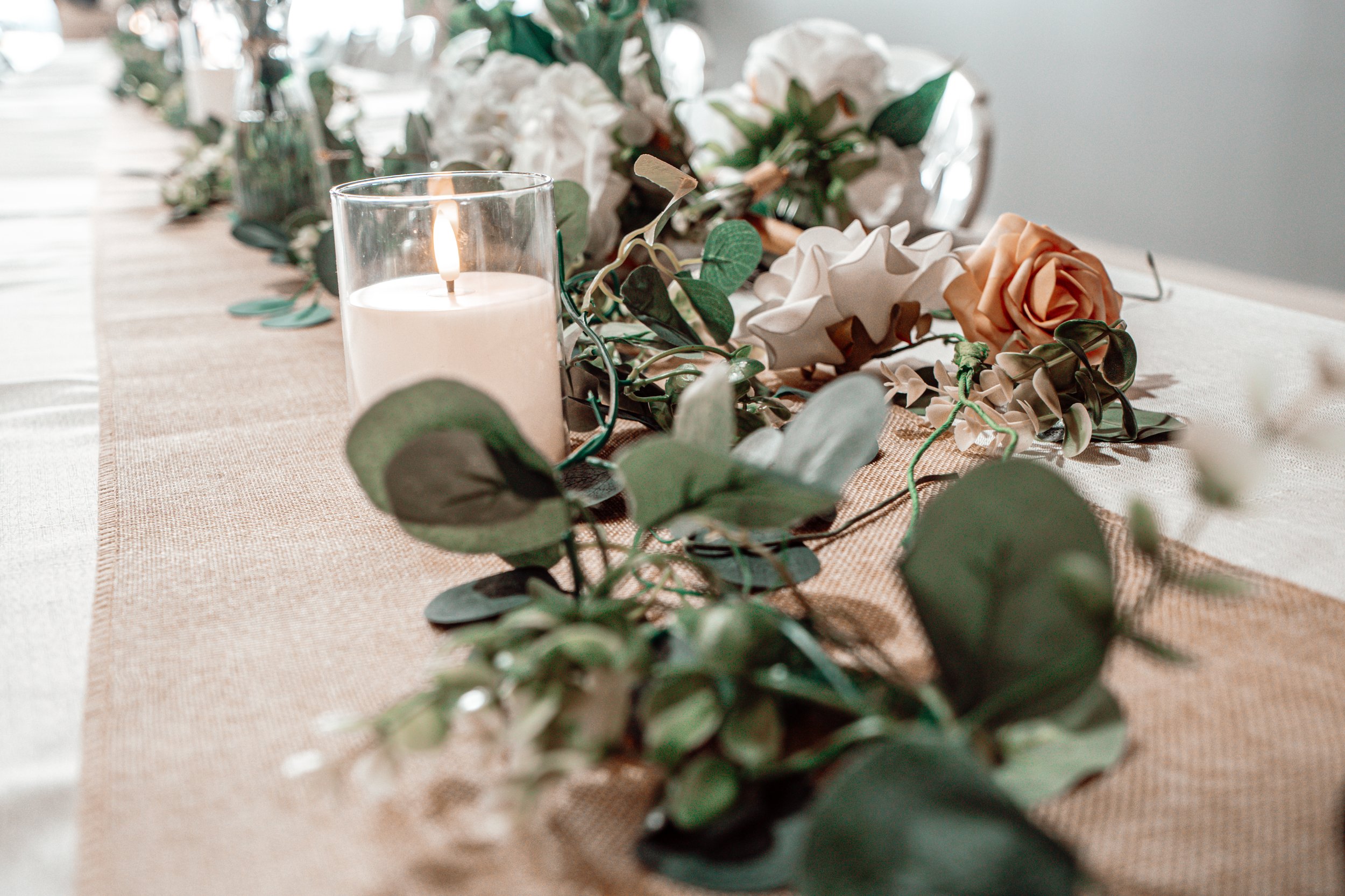 A lit white candle in a glass holder surrounded by a floral arrangement with white and peach roses, eucalyptus, and other greenery on a beige table runner.