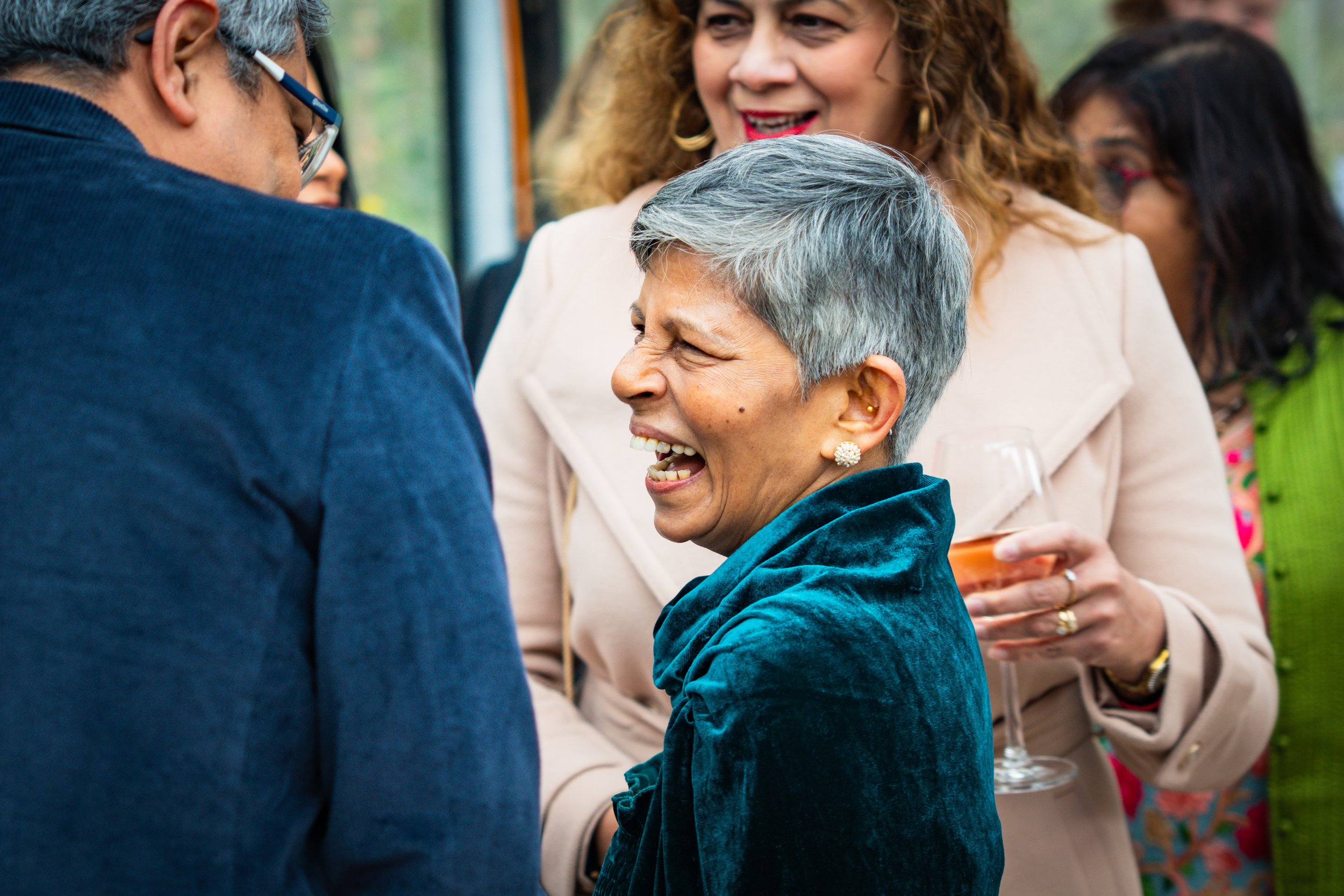 A group of people at a social gathering, smiling and engaging in conversation, with a woman in a teal outfit laughing in the center.