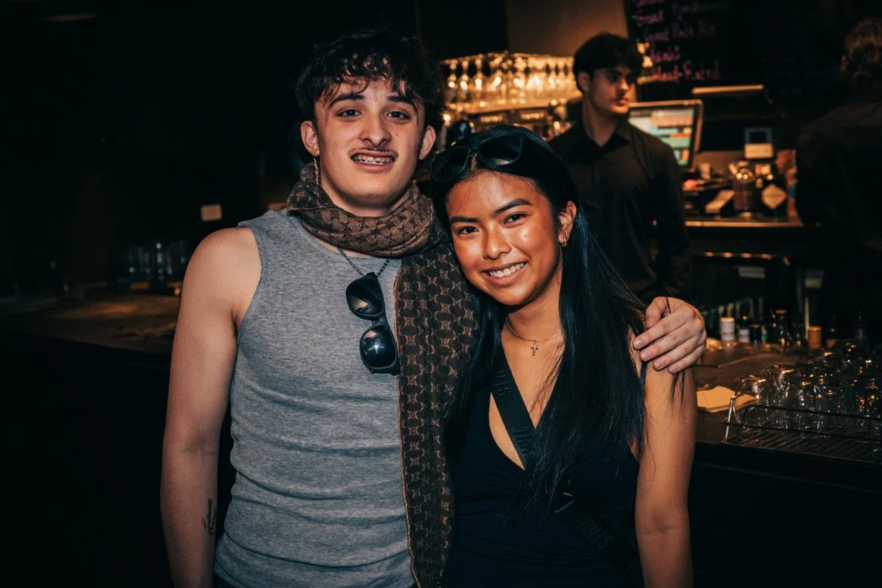 A young man and woman smiling in a bar or nightclub, with the man wearing a sleeveless shirt and scarf, and the woman wearing a black top and sunglasses on her head, standing close together with the man’s arm around the woman.