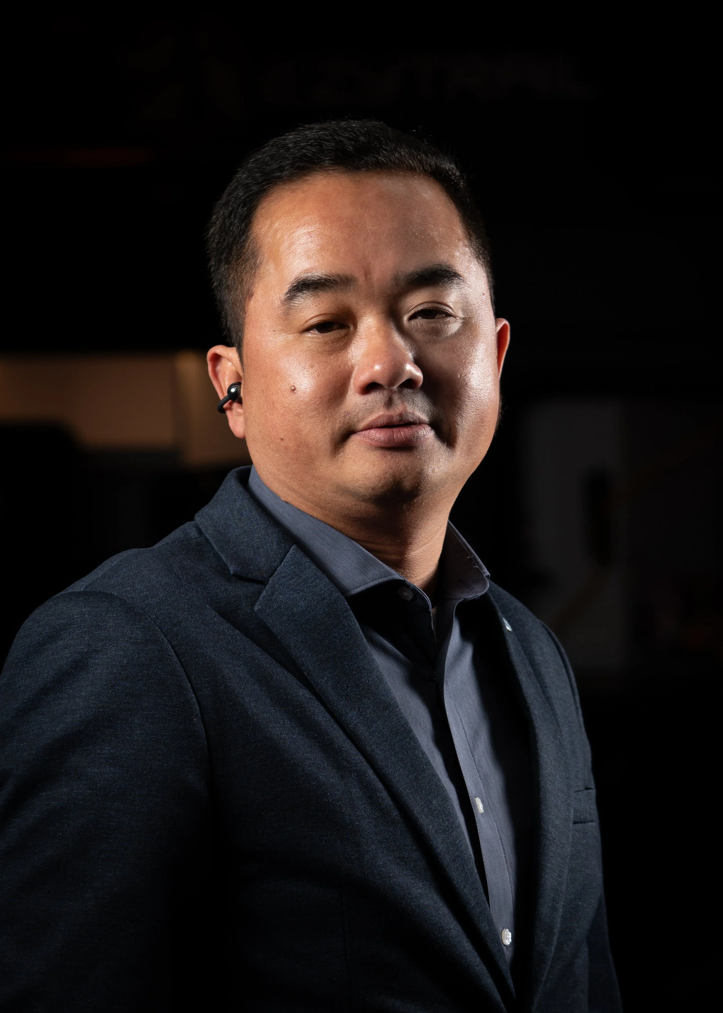 A man wearing a dark suit and shirt, with short black hair and a subtle smile, looking slightly to the side, set against a dark background.