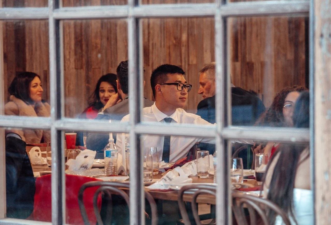 People sitting at a dinner table seen through a window with multiple panes, engaged in conversation.