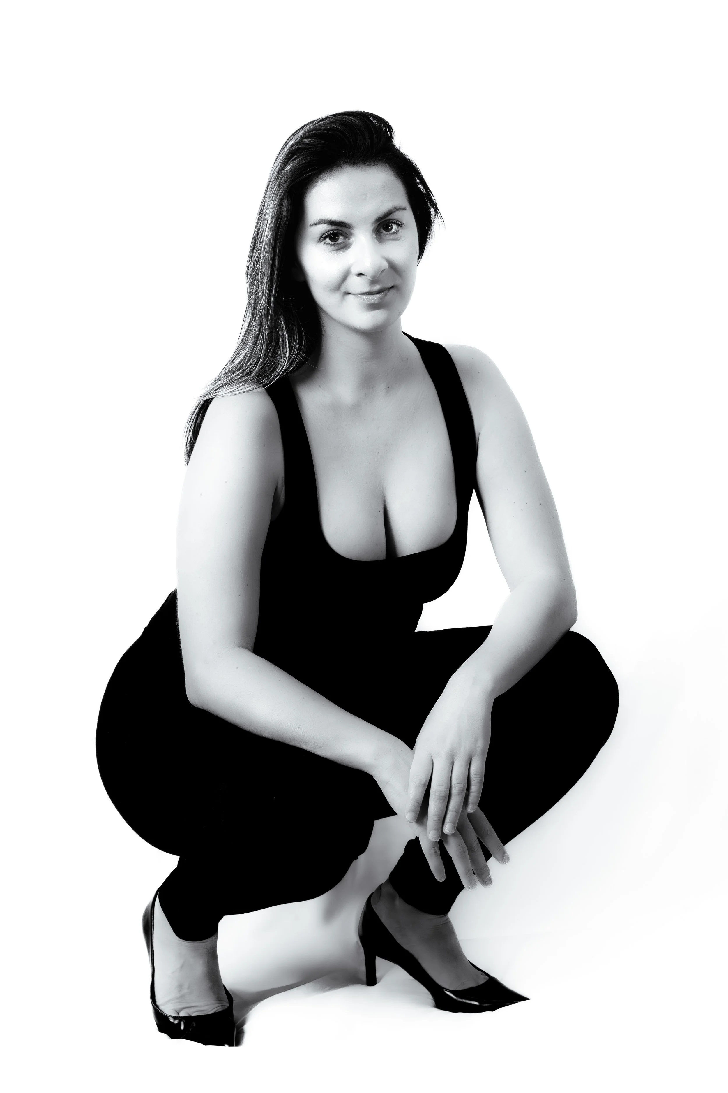 Black and white portrait of a woman with long dark hair, wearing a sleeveless top and high heel shoes, squatting against a plain background.