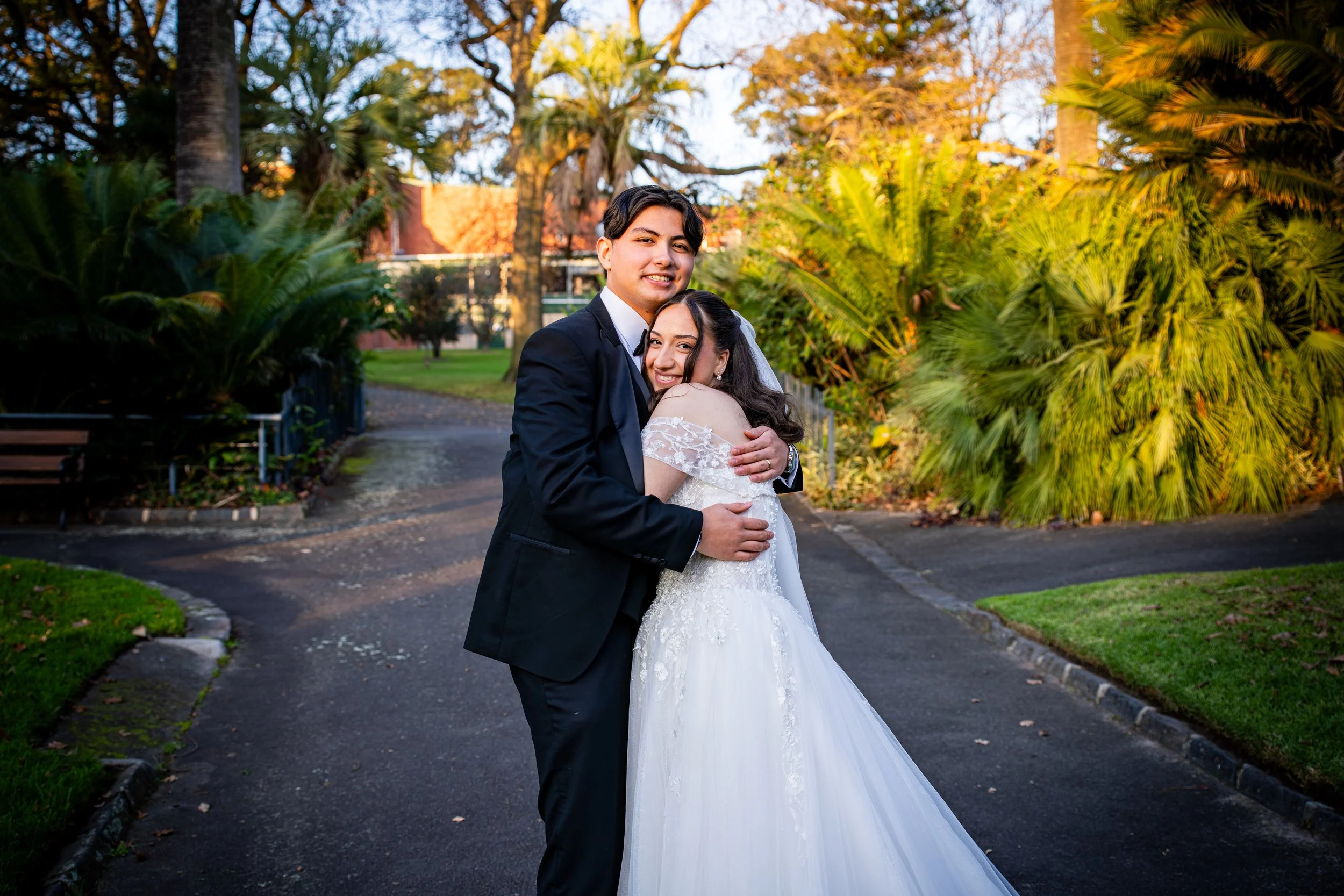 A newlywed couple dressed in wedding attire, embracing outdoors in a park with green plants and trees during late afternoon or early evening.
