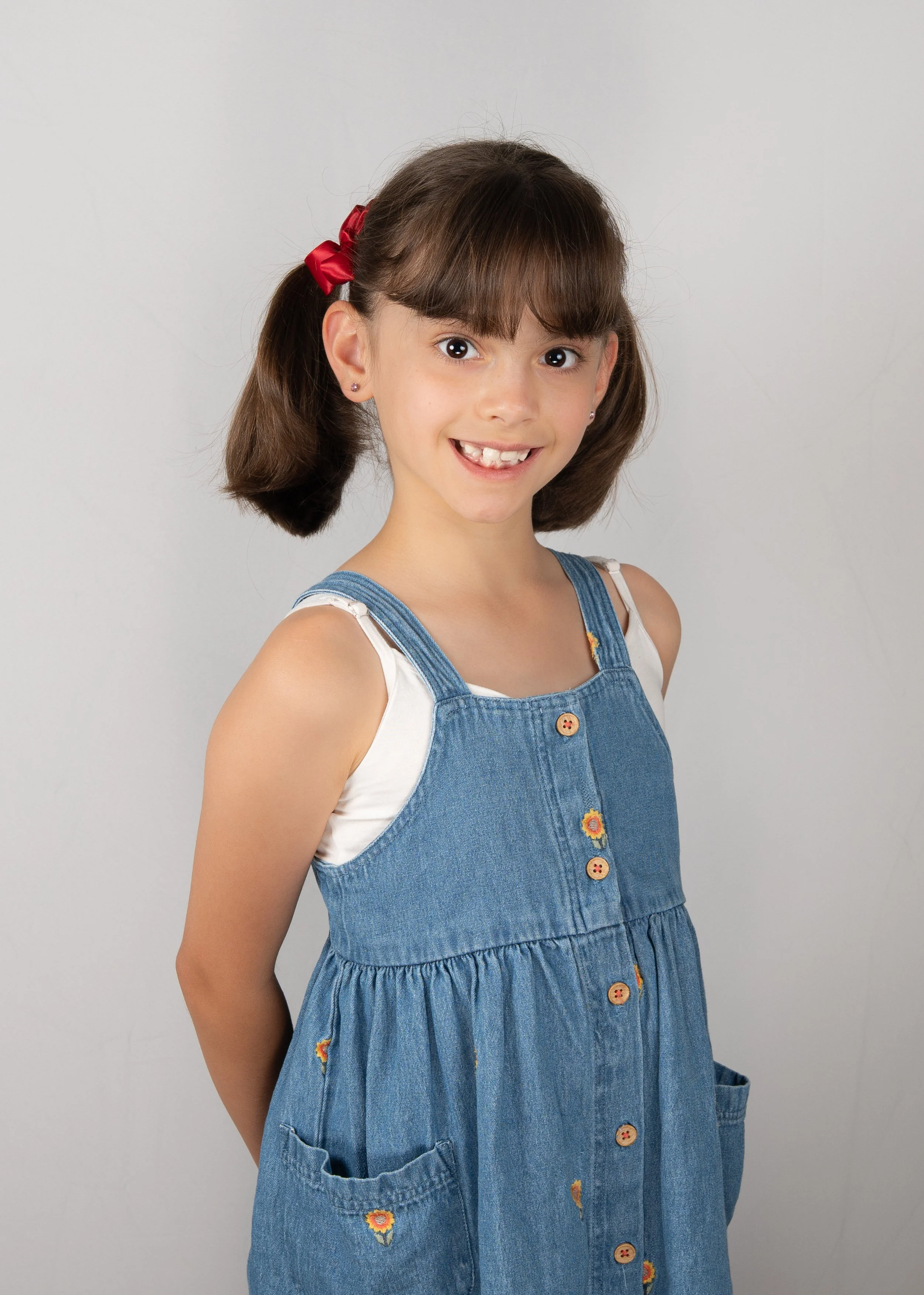 A young girl with brown hair styled in pigtails with a red ribbon, wearing a sleeveless white top and a denim dress with floral embroidery, smiling at the camera.