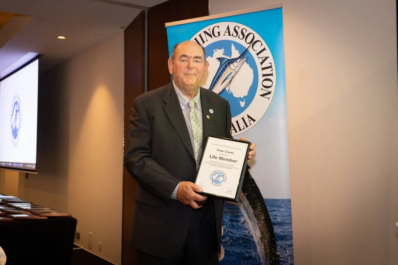 An older man in a suit holding a framed certificate stands in front of a banner with a fish and a map of Australia, at an event organized by the Fishing Association Australia.