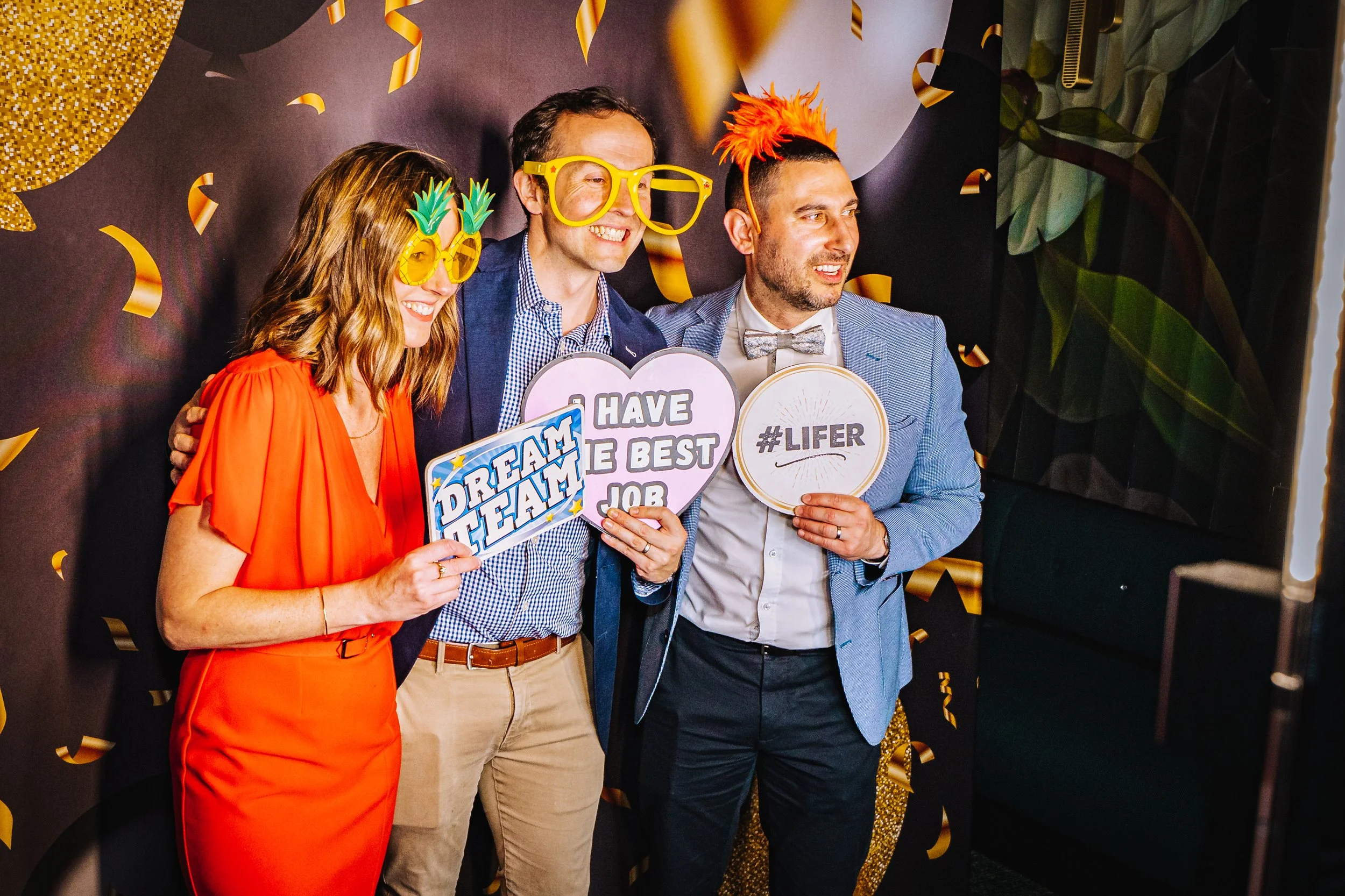 Three people at a celebration event wearing festive glasses and holding signs with positive messages. The background is decorated with black and gold streamers and balloons.