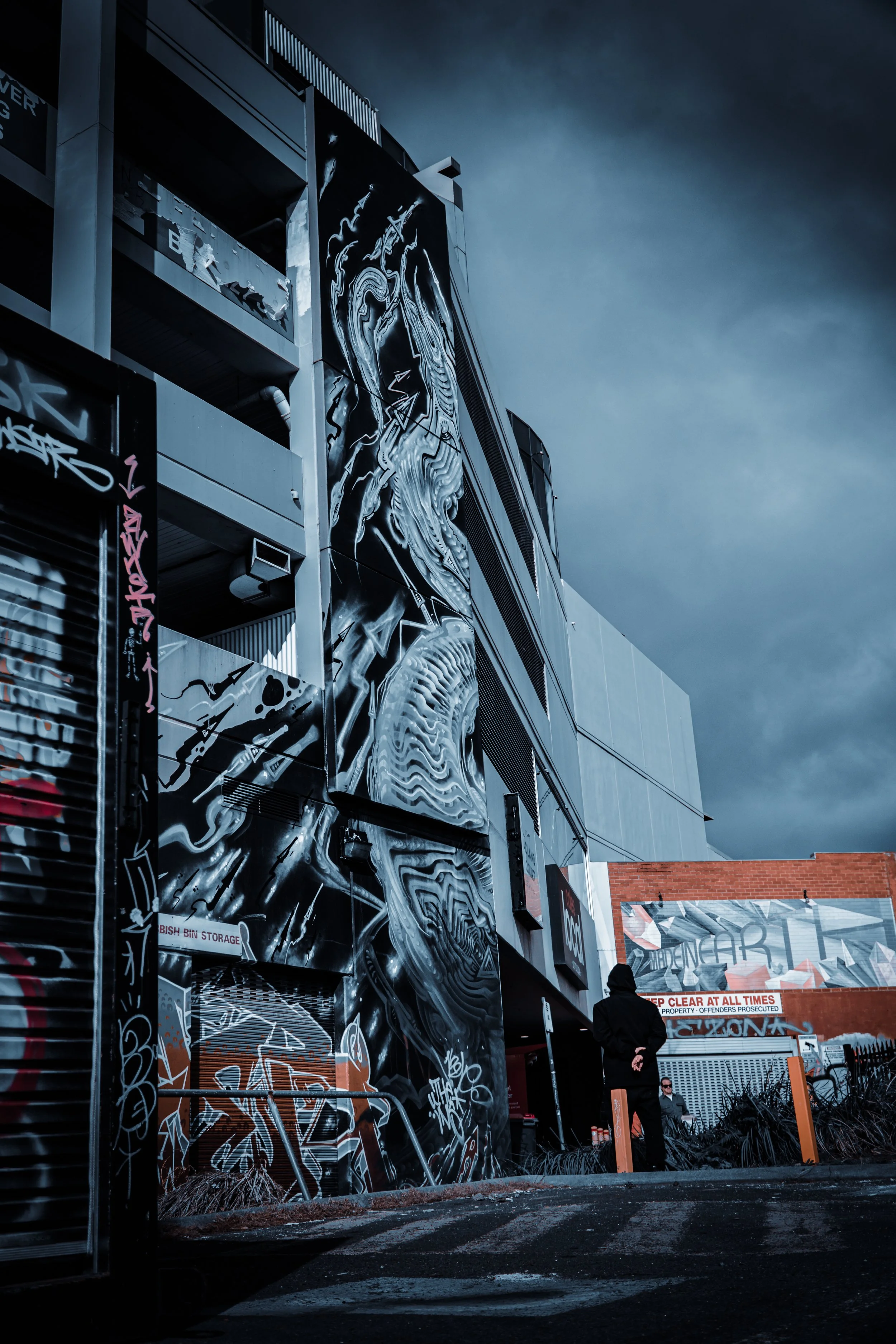 Urban scene featuring a large building with vibrant graffiti art, a person in a hoodie standing nearby, and a cloudy sky overhead.
