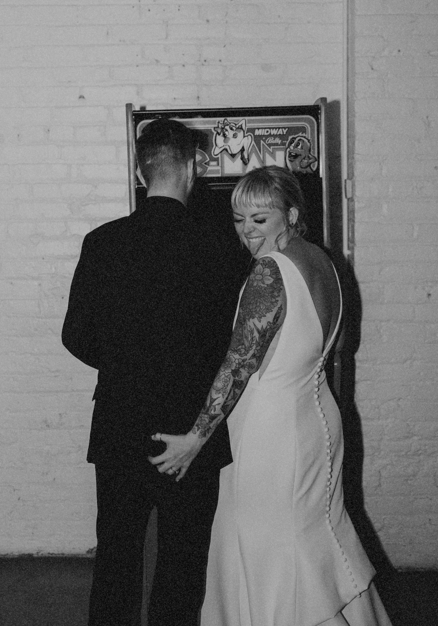A woman in a white dress with tattoos on her arm holding hands with a man in a dark suit, standing in front of a video game machine, making a playful face with her tongue out.