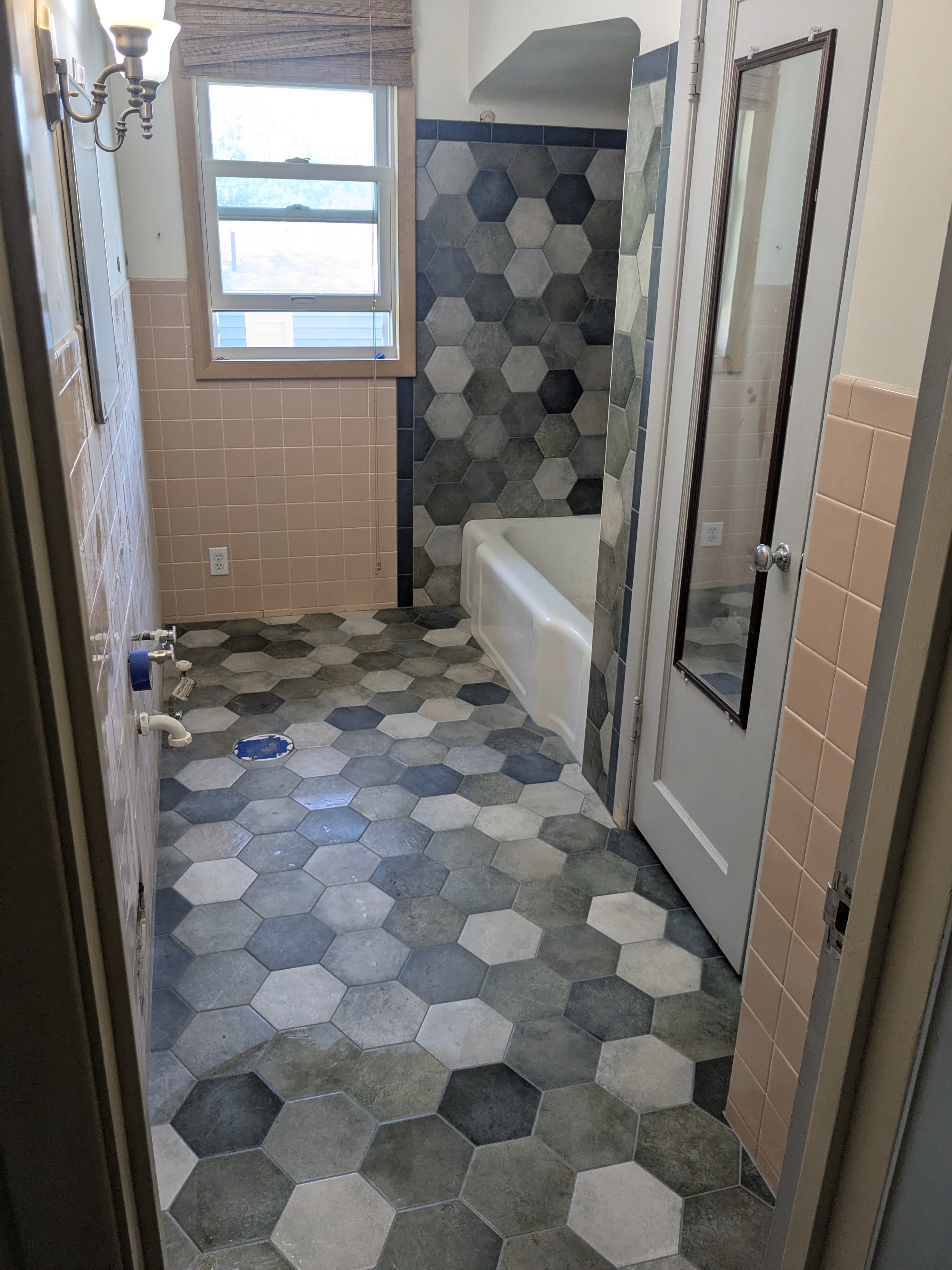 A bathroom under renovation with hexagonal tile floor in shades of gray, a wall with pink tiles, a window, a bathtub, and a door with a mirror.