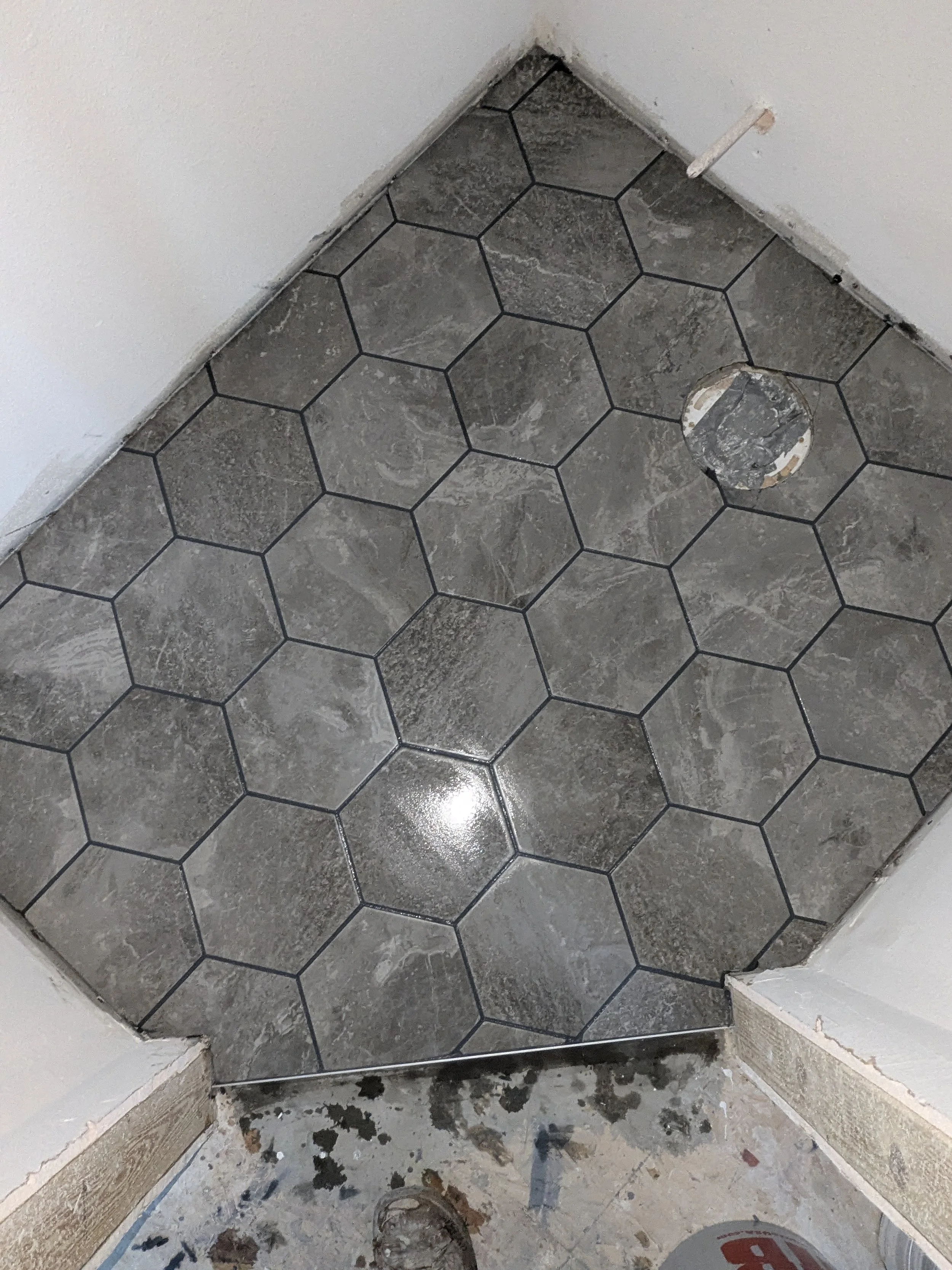Bathroom floor with large, hexagonal gray tiles, one tile with a hole and missing grout, partially installed transition strip at the entry, and construction debris on the floor.