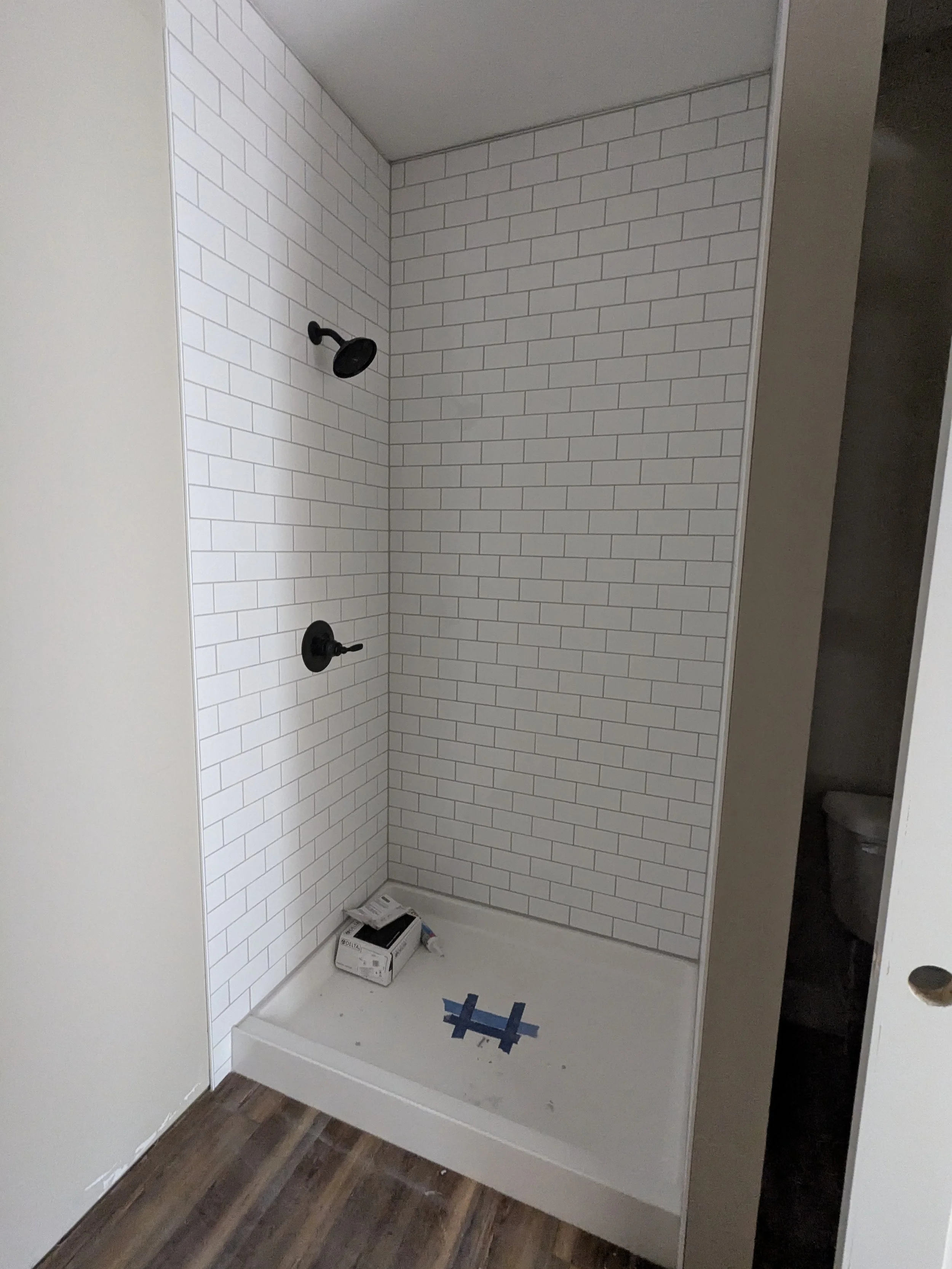 Bathroom shower with white subway tile walls, black shower fixtures, and an unfinished base with a box and blue tape on the floor.