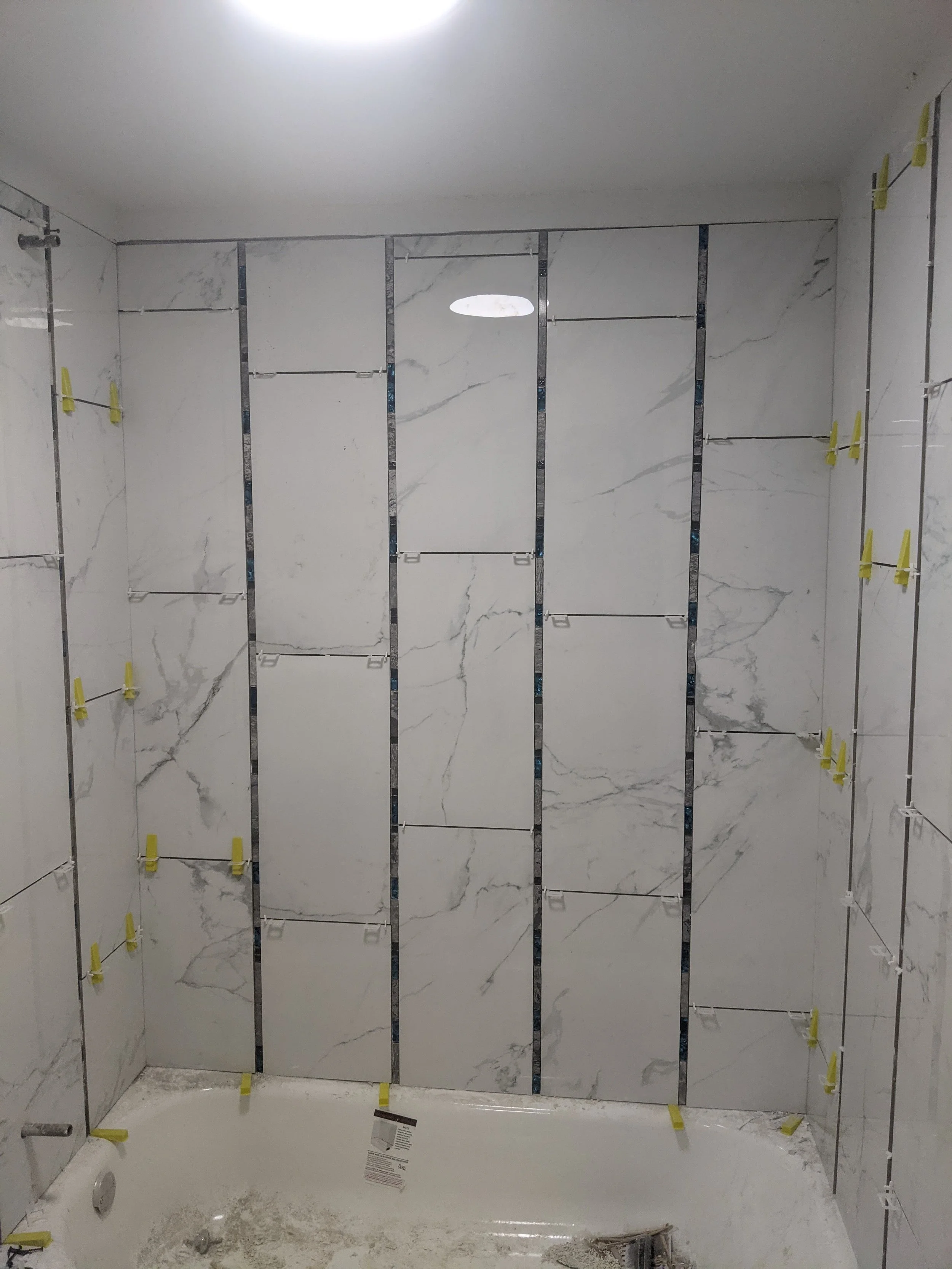 Bathroom with marble wall tiles under installation, with yellow clips holding tiles in place, a shower drain and partially finished floor.
