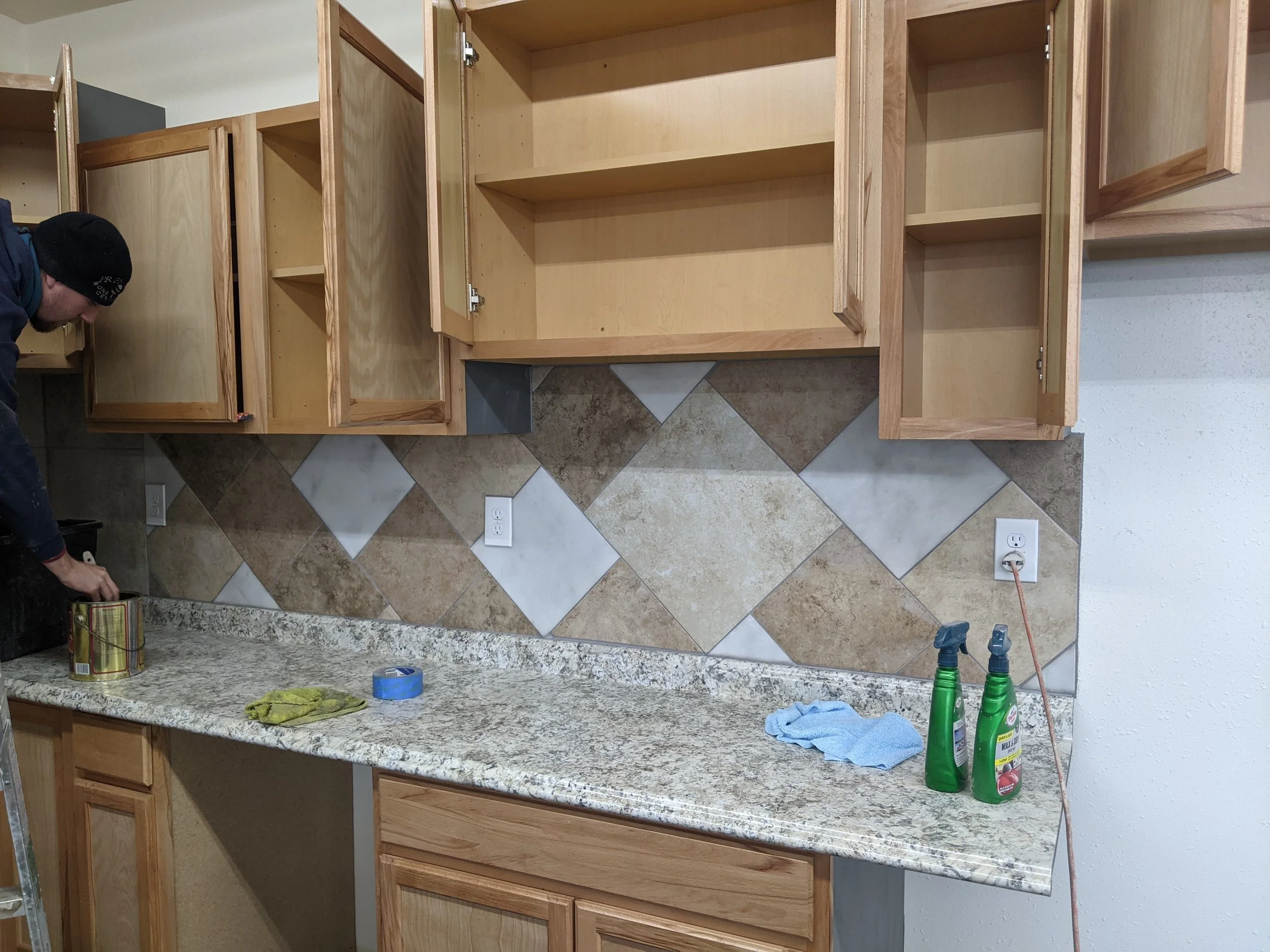A kitchen undergoing renovation with open wooden cabinets, a granite countertop, tile backsplash, and cleaning supplies on the counter. A person is partially visible on the left side.