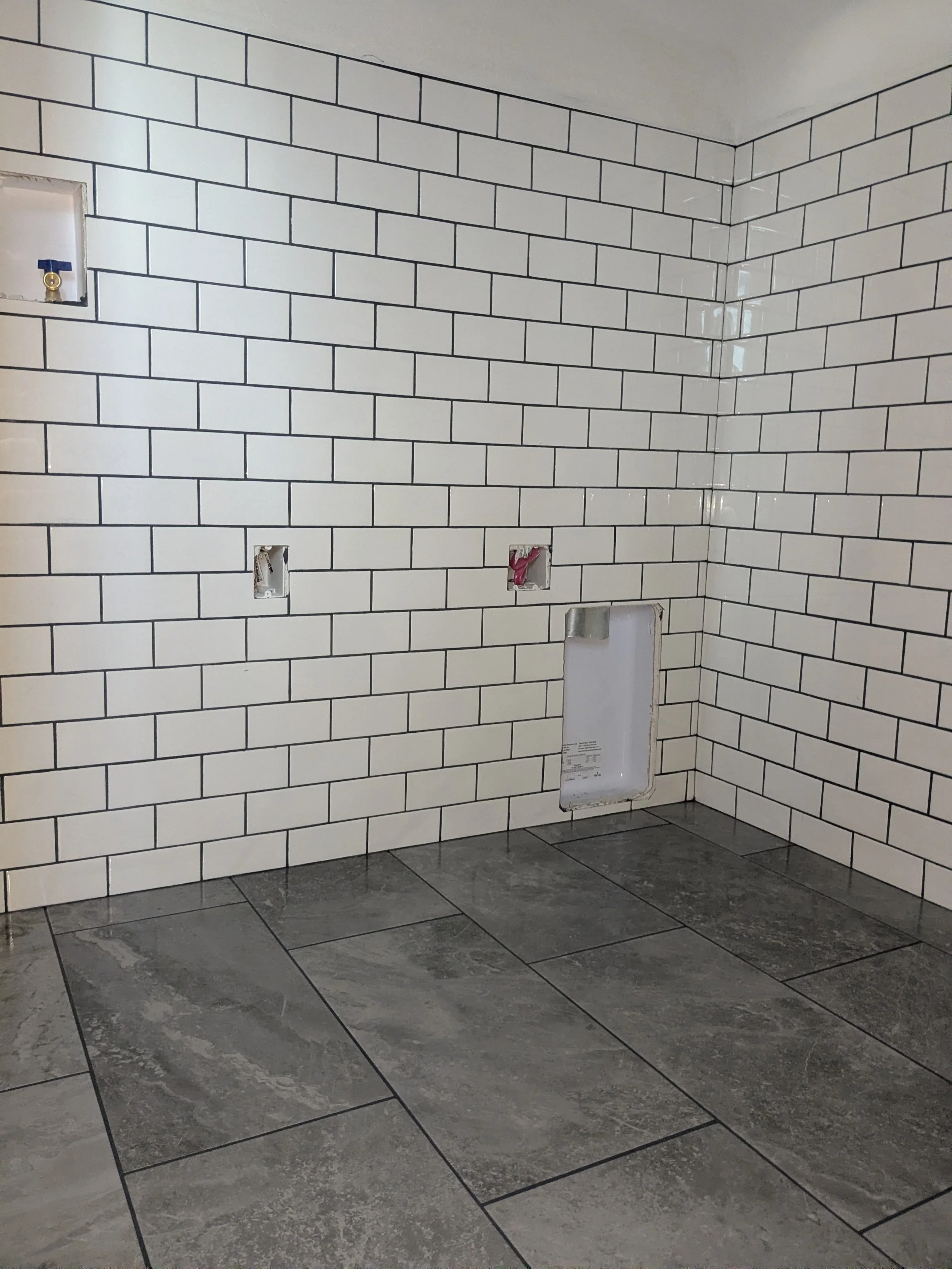 White subway tile wall with three exposed wall outlets and a square opening, gray tiled floor in a room under construction or renovation.