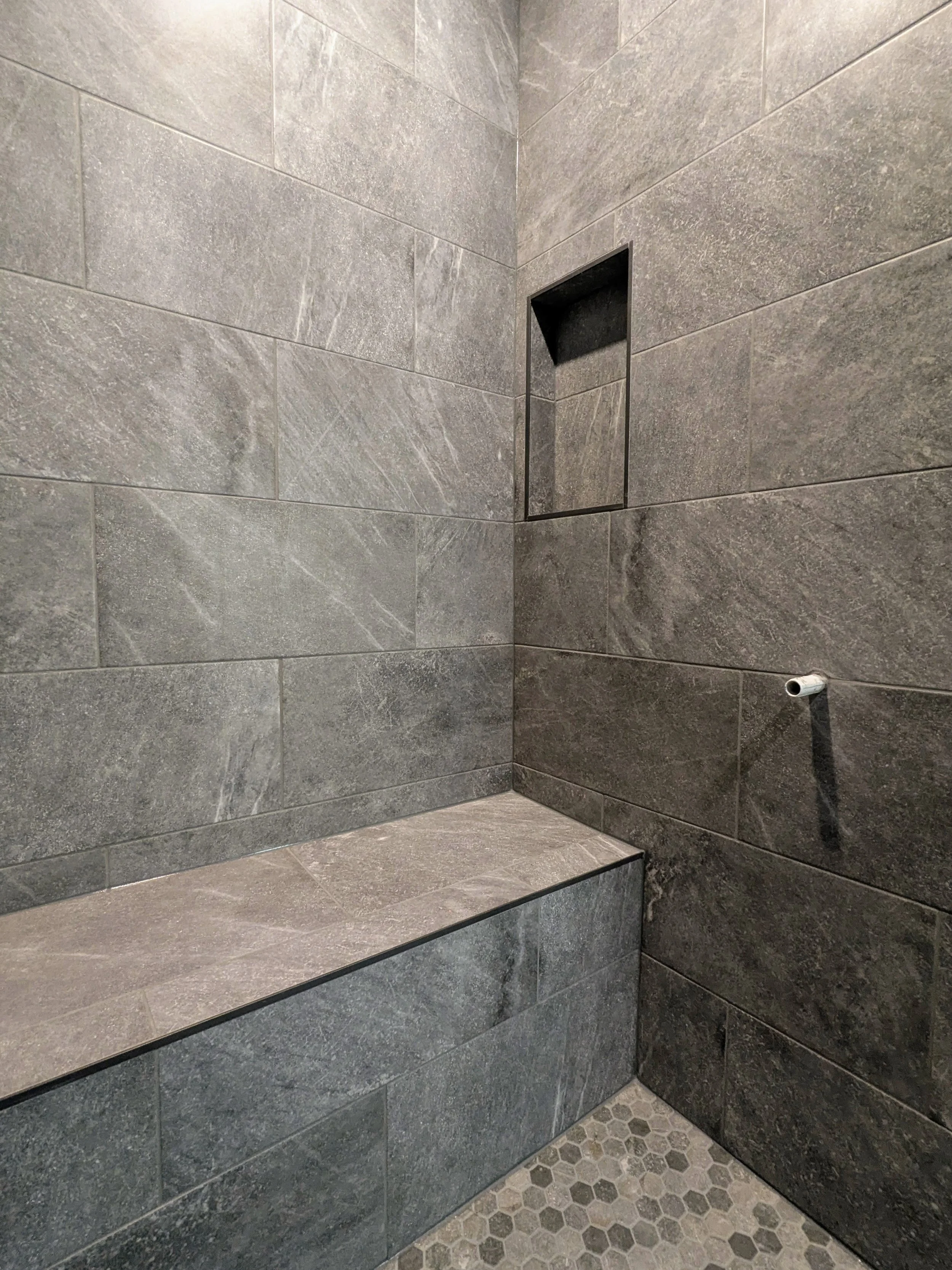 Corner of a shower with gray tile walls, a built-in shelf, and hexagon tile flooring.