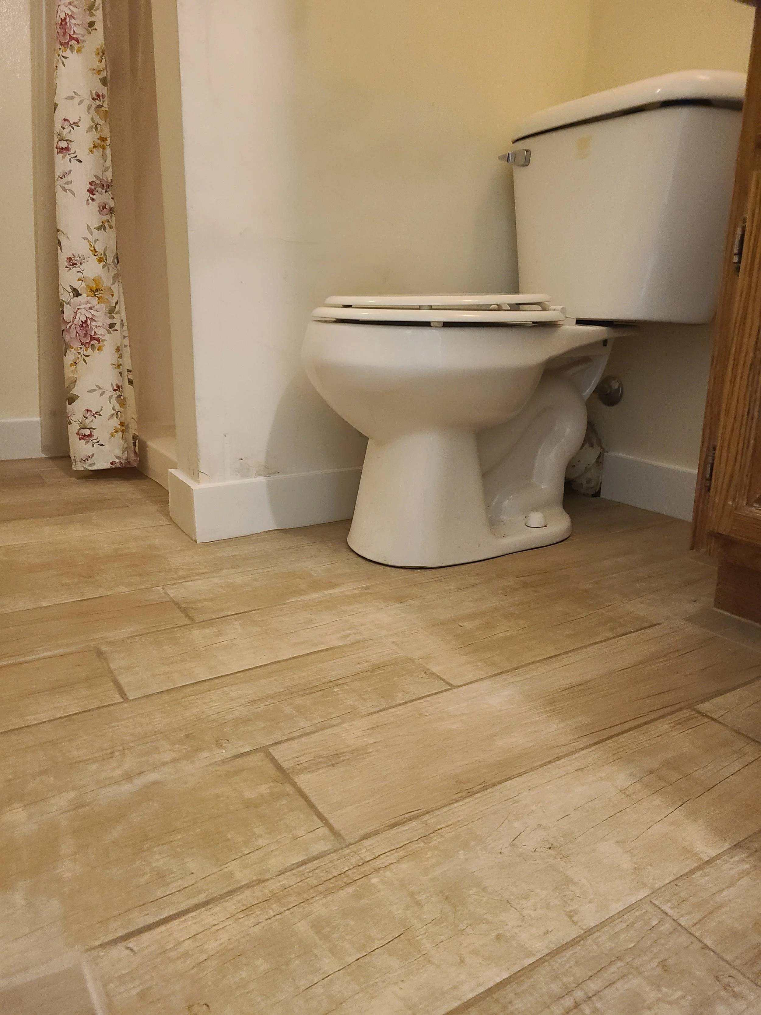 A close-up photo of a white toilet with the lid open in a bathroom, next to a wooden cabinet and a floral curtain.