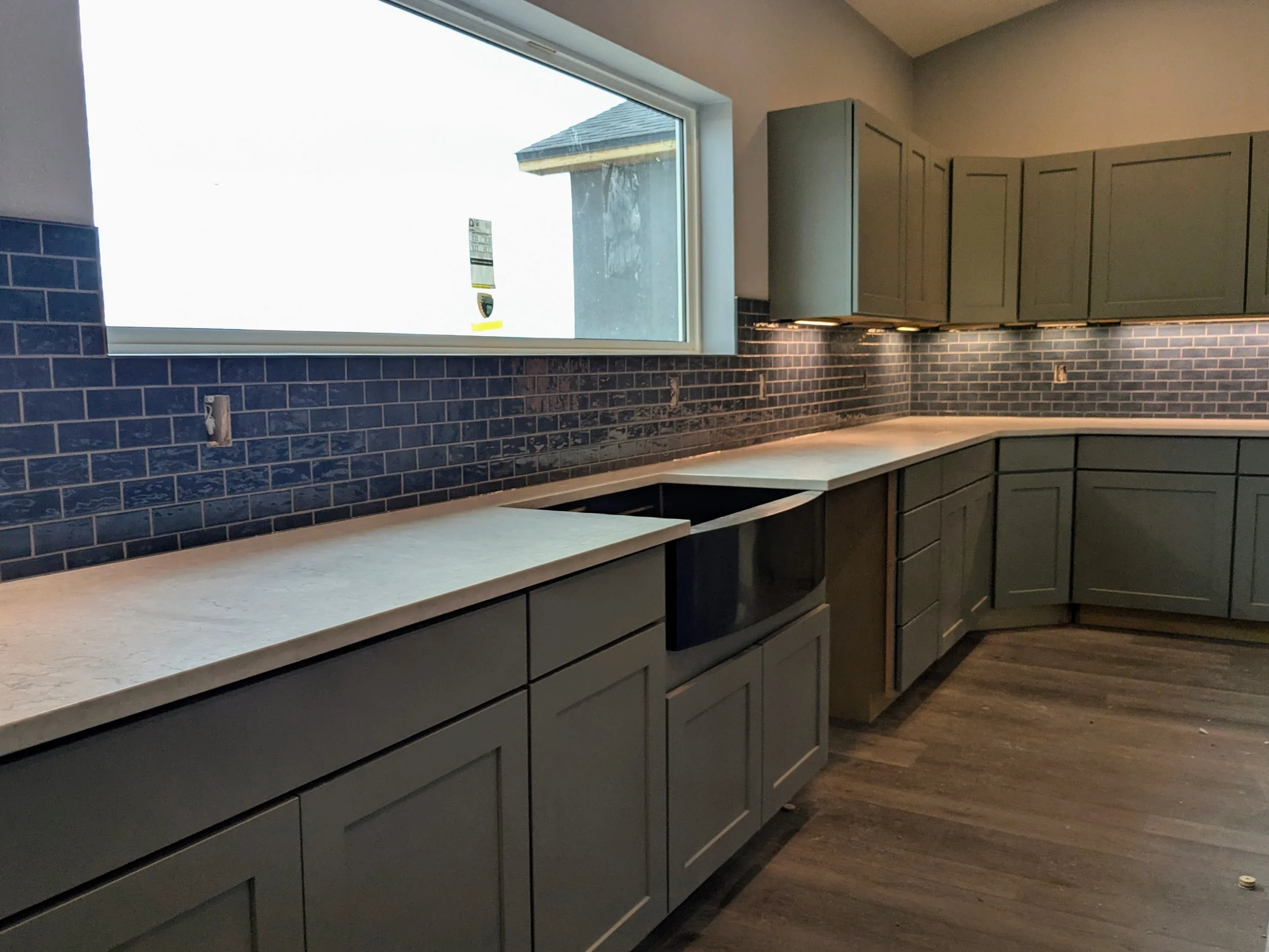 Empty kitchen with green cabinets, a black microwave, blue subway tile backsplash, and a large window with a view of the exterior house wall.