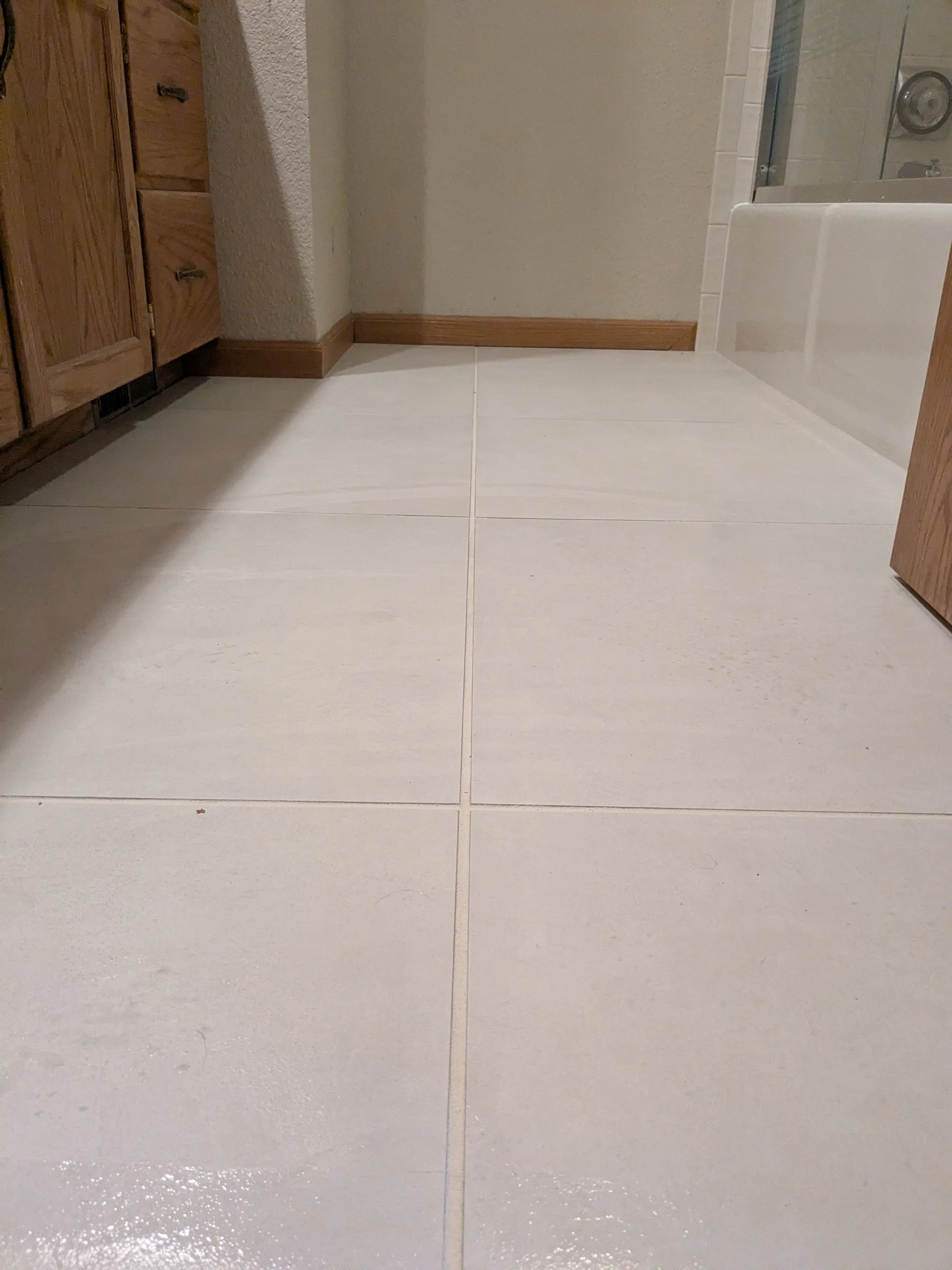 Bathroom floor with large white tiles, wooden cabinets on the left, a partial view of a bathtub on the right, and a textured beige wall in the background.
