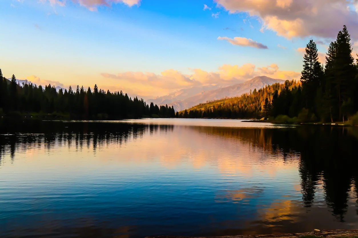 A serene lake surrounded by pine trees with mountains in the background during sunrise, reflecting colorful clouds in the water.