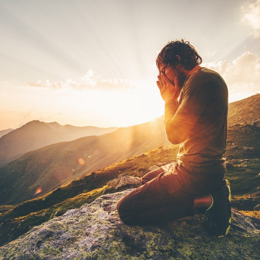 A person kneeling on a rock on a mountain during sunrise, holding their face with both hands, appearing to pray or meditate, with scenic mountains and a partly cloudy sky in the background.