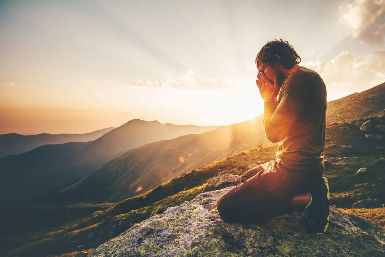 Person kneeling on a rock with hands clasped in prayer or reflection, overlooking mountains during sunrise.