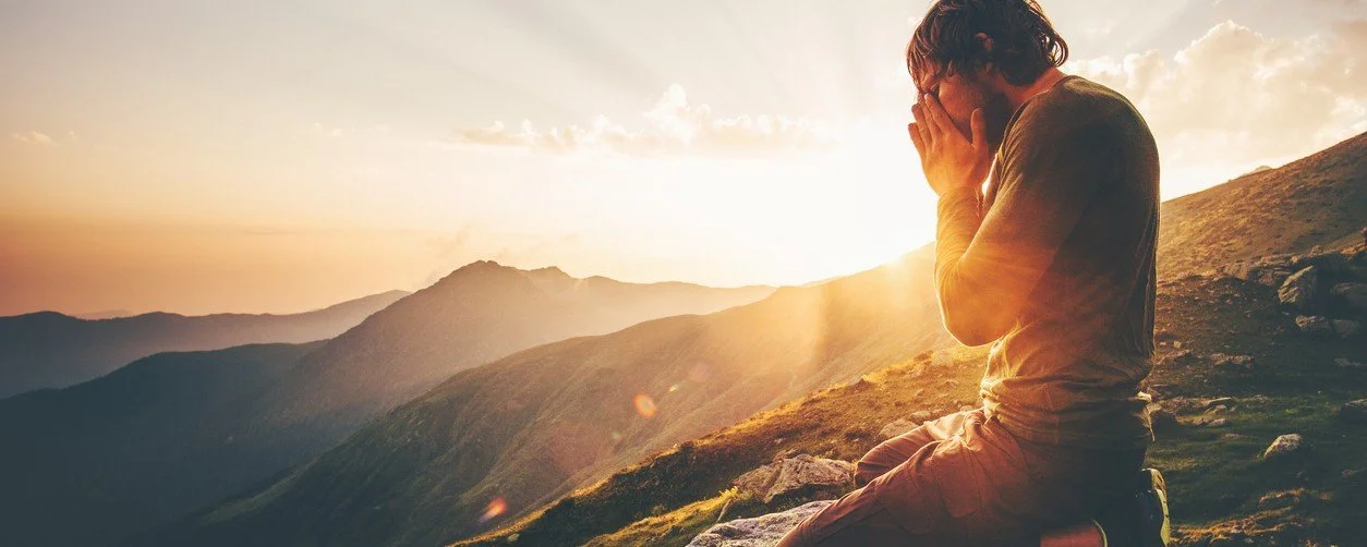 Young man kneeling on mountain ridge at sunrise, with with his praying hands caressing his face.