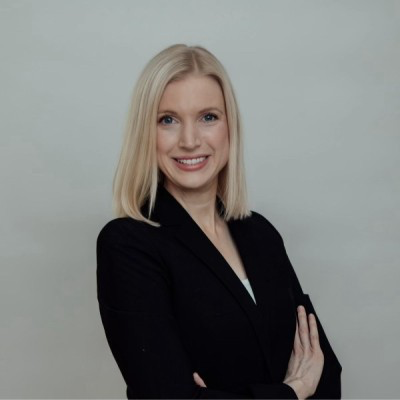 A woman with blonde hair wearing a black blazer, smiling with arms crossed, against a plain light gray background.
