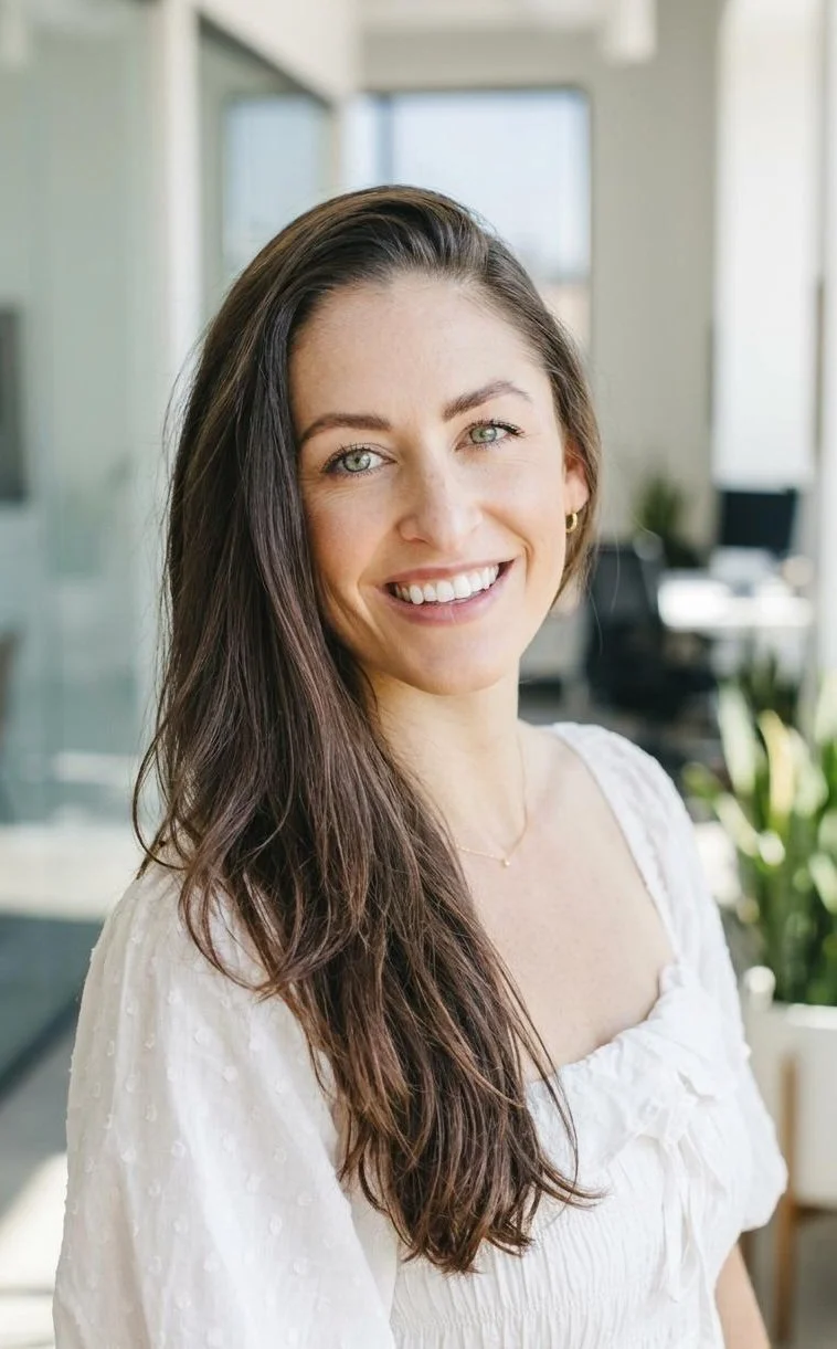A woman with long brown hair and green eyes smiling, standing indoors near large windows with a blurred background of office furniture and plants.