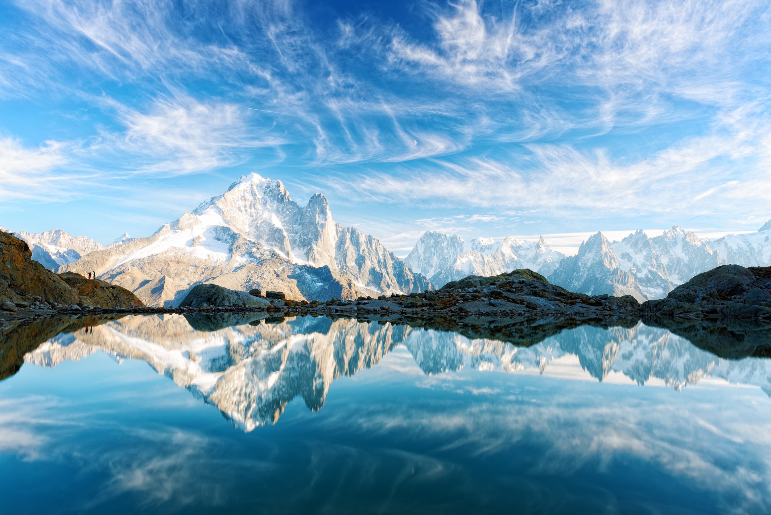 Snow-capped mountains reflecting in a calm mountain lake beneath a blue sky with wispy clouds.