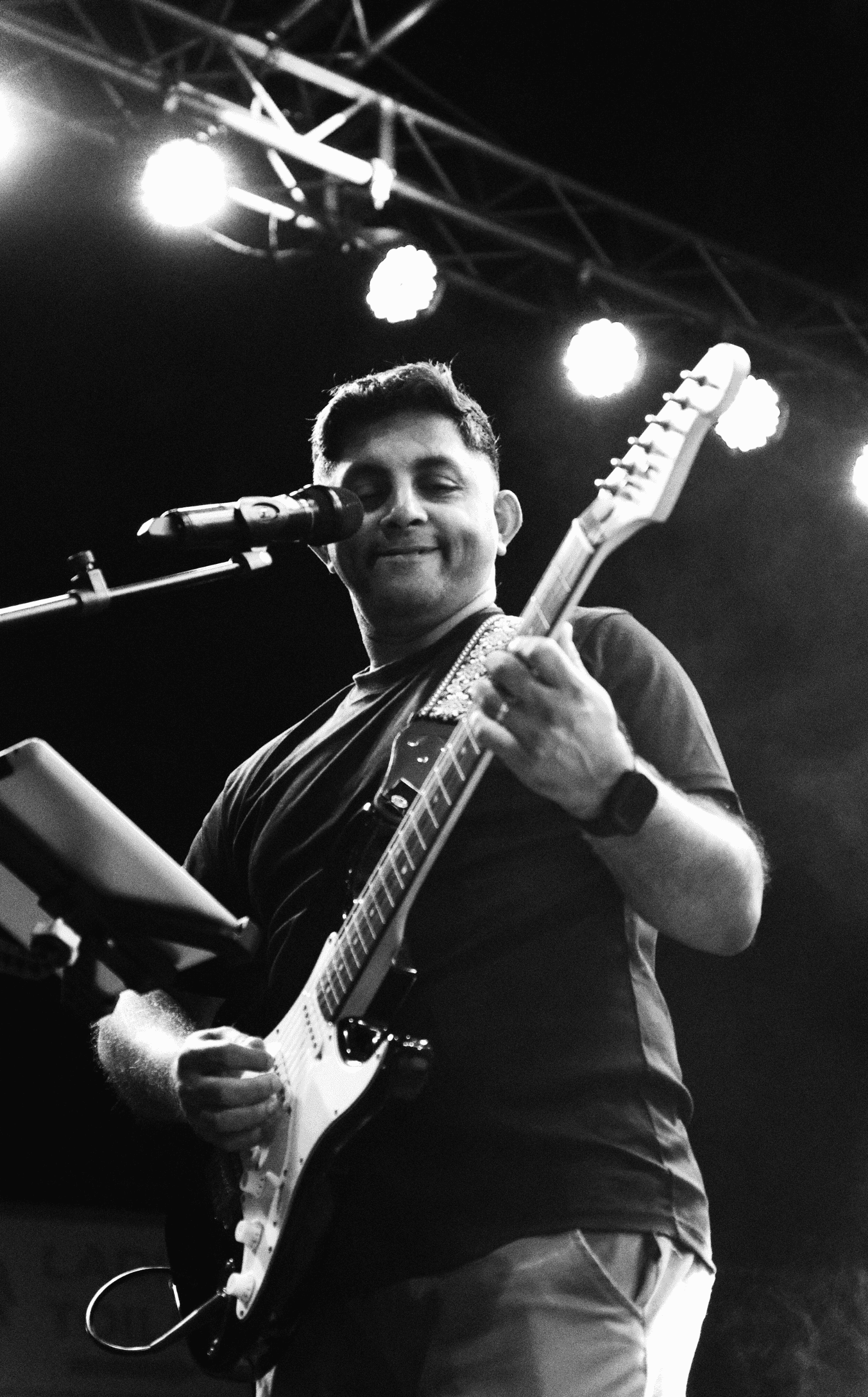 A man playing an electric guitar on stage, smiling, with stage lights above him.