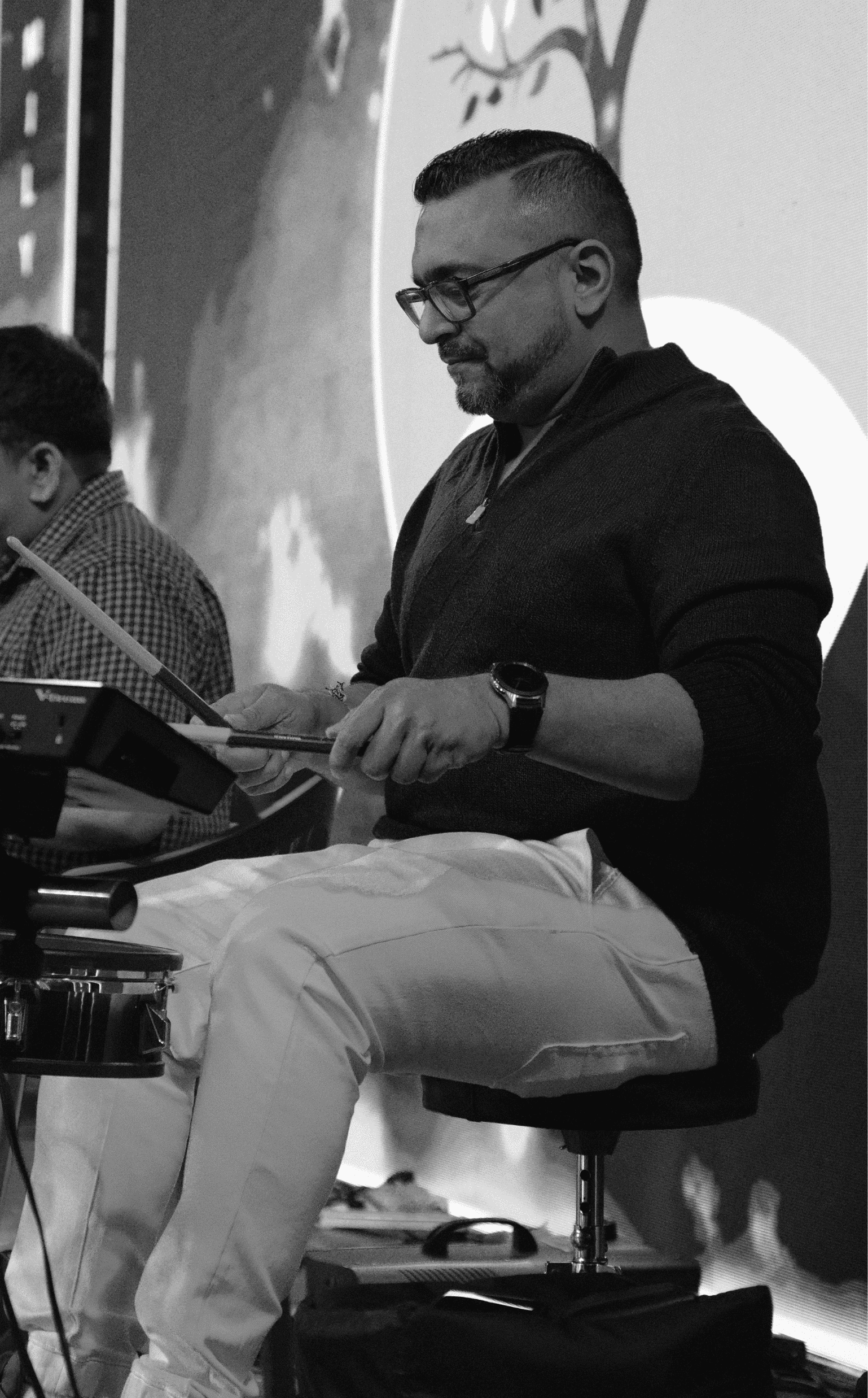 A man wearing glasses, a dark sweater, and a watch, sitting on a stool and looking at a mobile device, with another person partially visible beside him, in a room with a large circular graphic on the wall behind them.