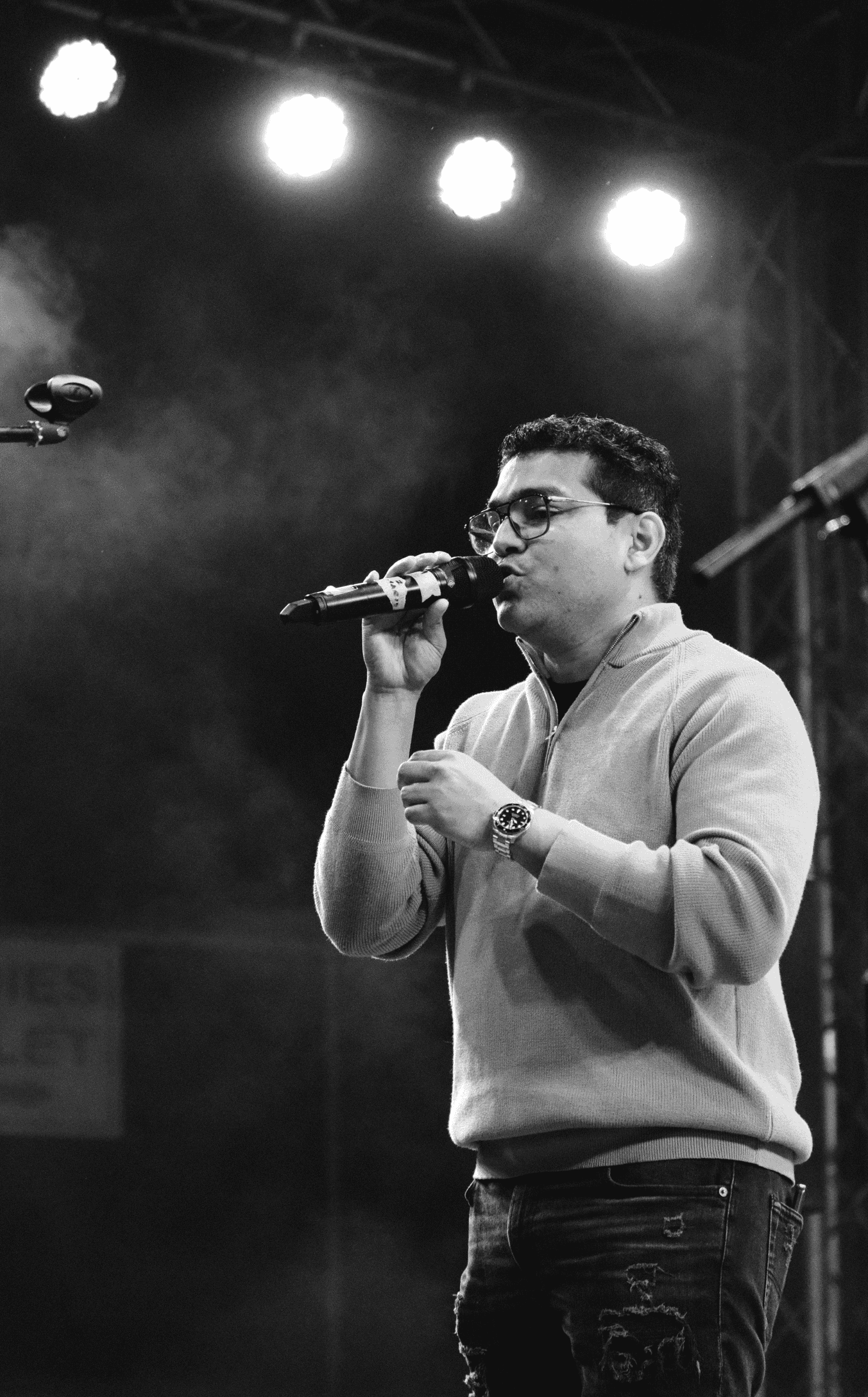 A man wearing glasses and a light-colored sweater is singing into a microphone on stage, illuminated by bright stage lights overhead, in a black-and-white photo.